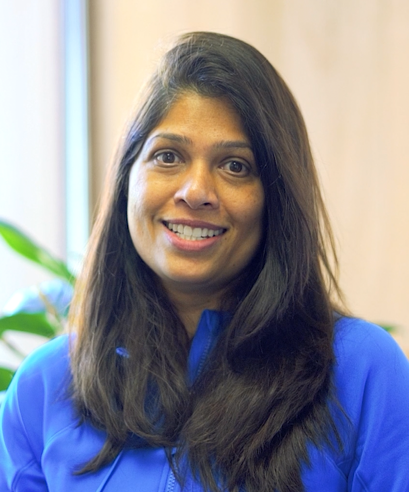 A woman with long dark hair smiling, wearing a blue jacket, standing indoors near a window with a plant in the background.