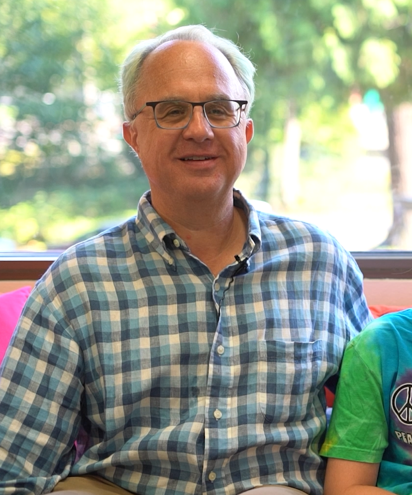 A smiling middle-aged man with light-colored hair and glasses, wearing a blue plaid shirt, sitting indoors with a window showing greenery outside.