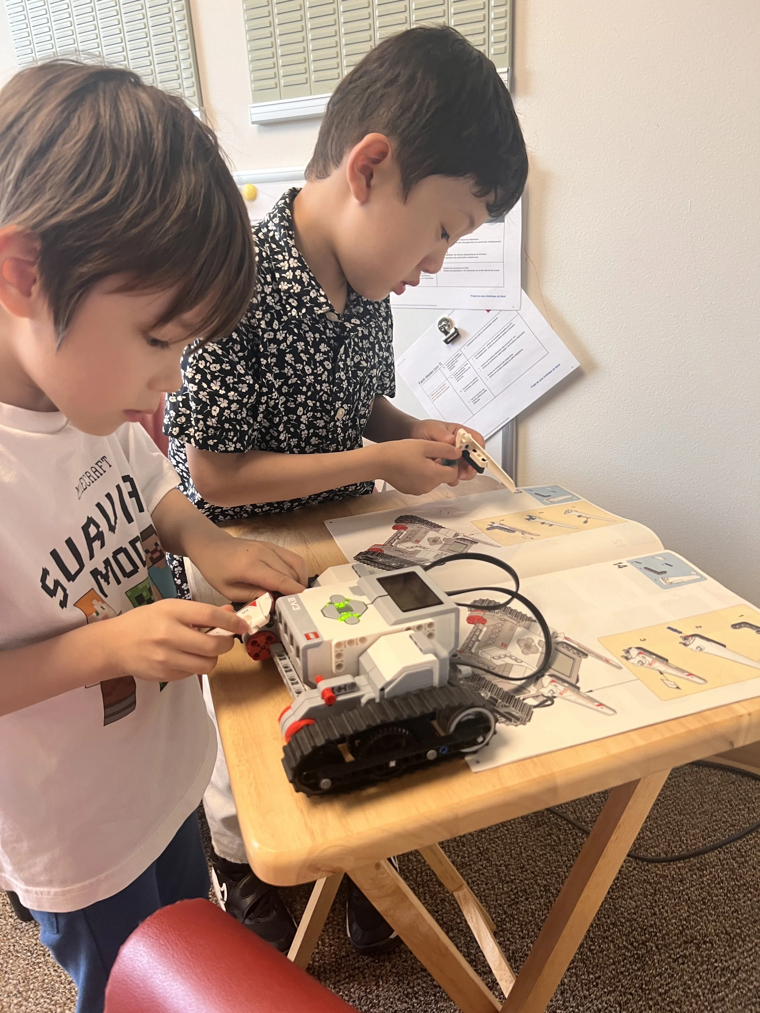 Two boys working on building a LEGO robot at a table, with instruction manuals and a laptop nearby.