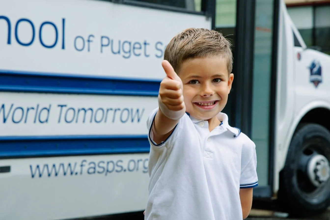 A smiling young boy giving a thumbs up in front of a FASPS school bus.
