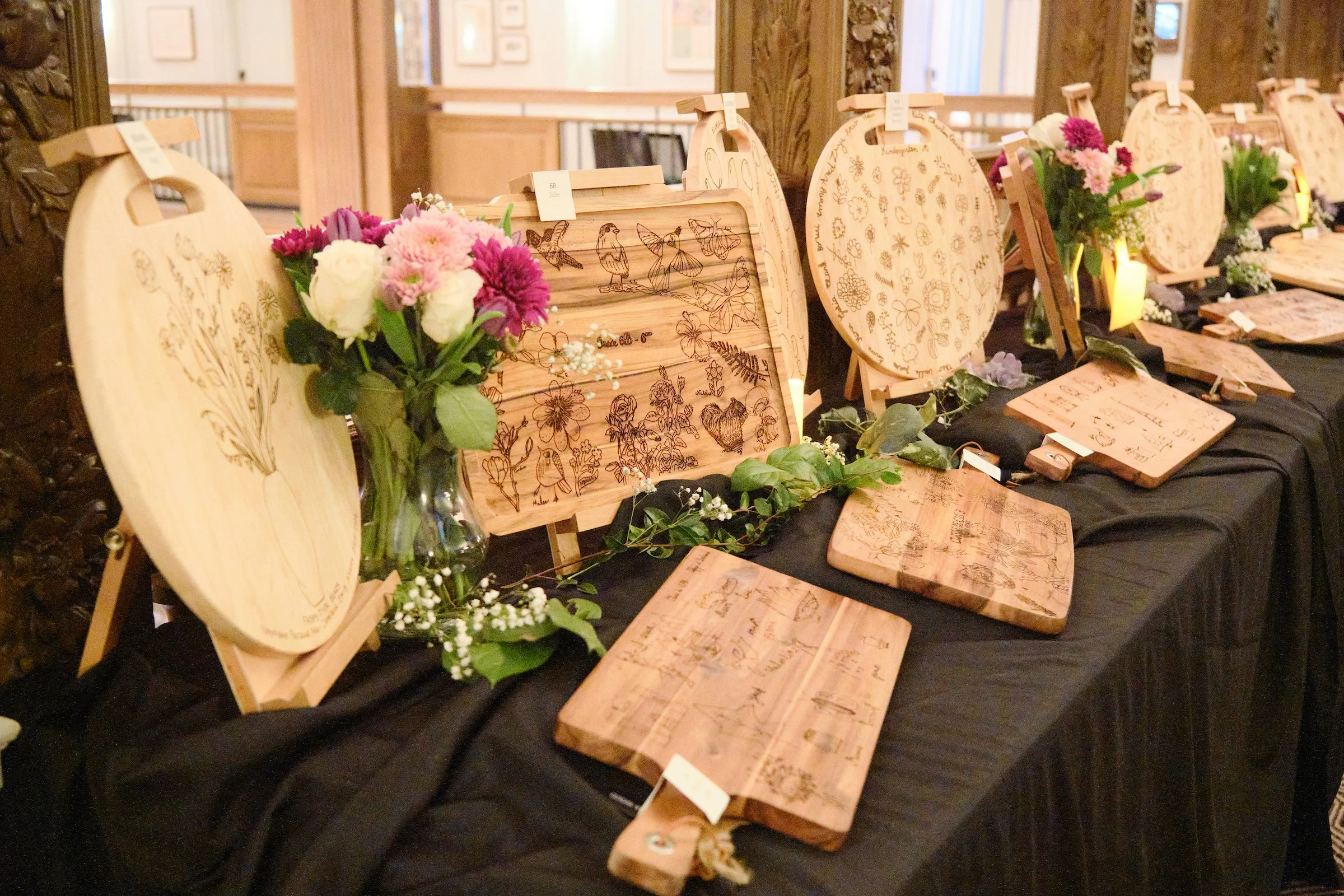 Display of wood engraved items, including cutting boards and trays, with floral and butterfly designs, accompanied by pink, white, and purple flowers, on a black cloth surface in a well-lit room.