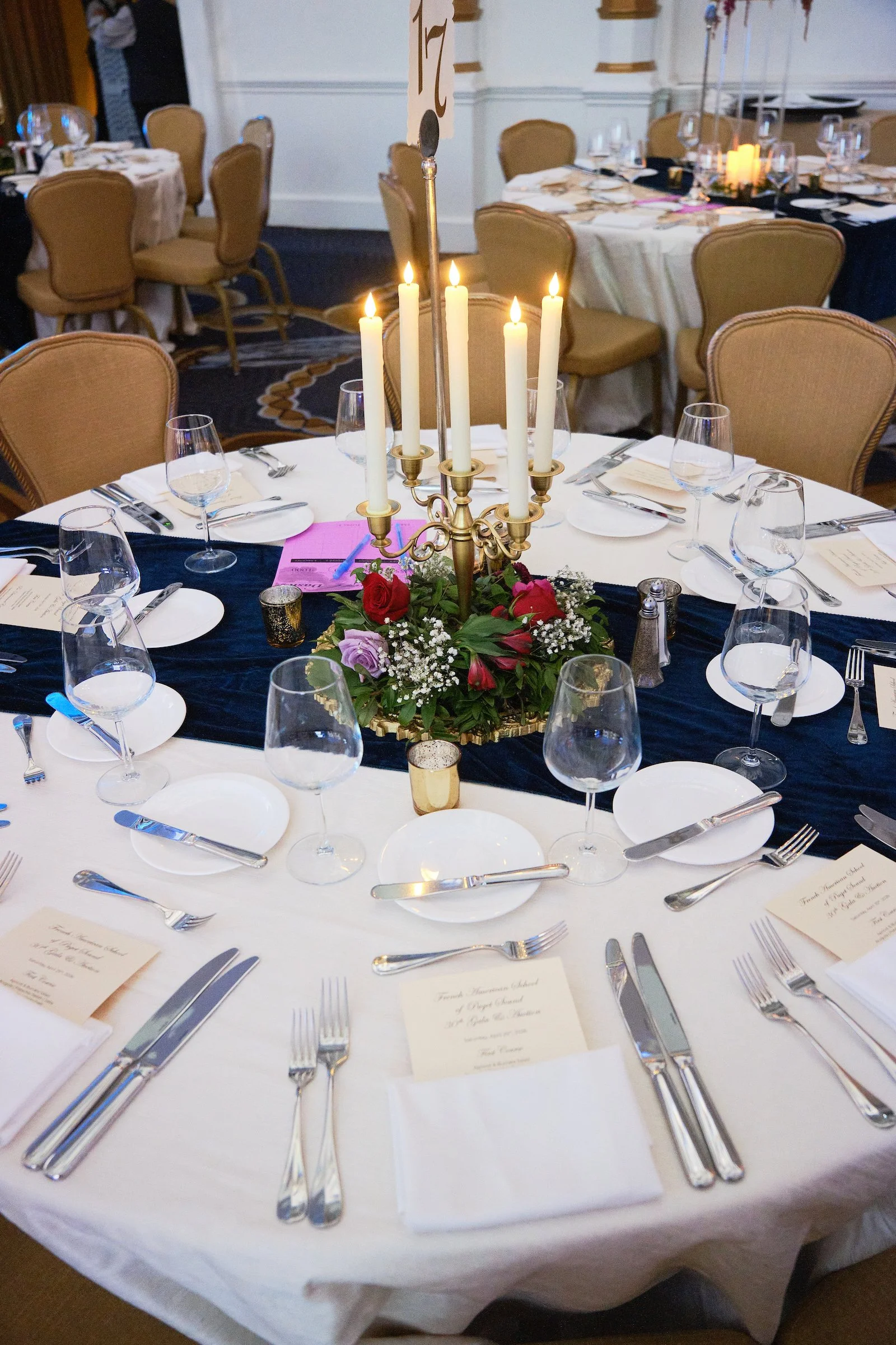 Round banquet table set for a formal dinner with white tablecloth, arranged silverware, wine glasses, and folded napkins, featuring a centerpiece with lit candles and floral decorations. Multiple similar tables can be seen in the background.