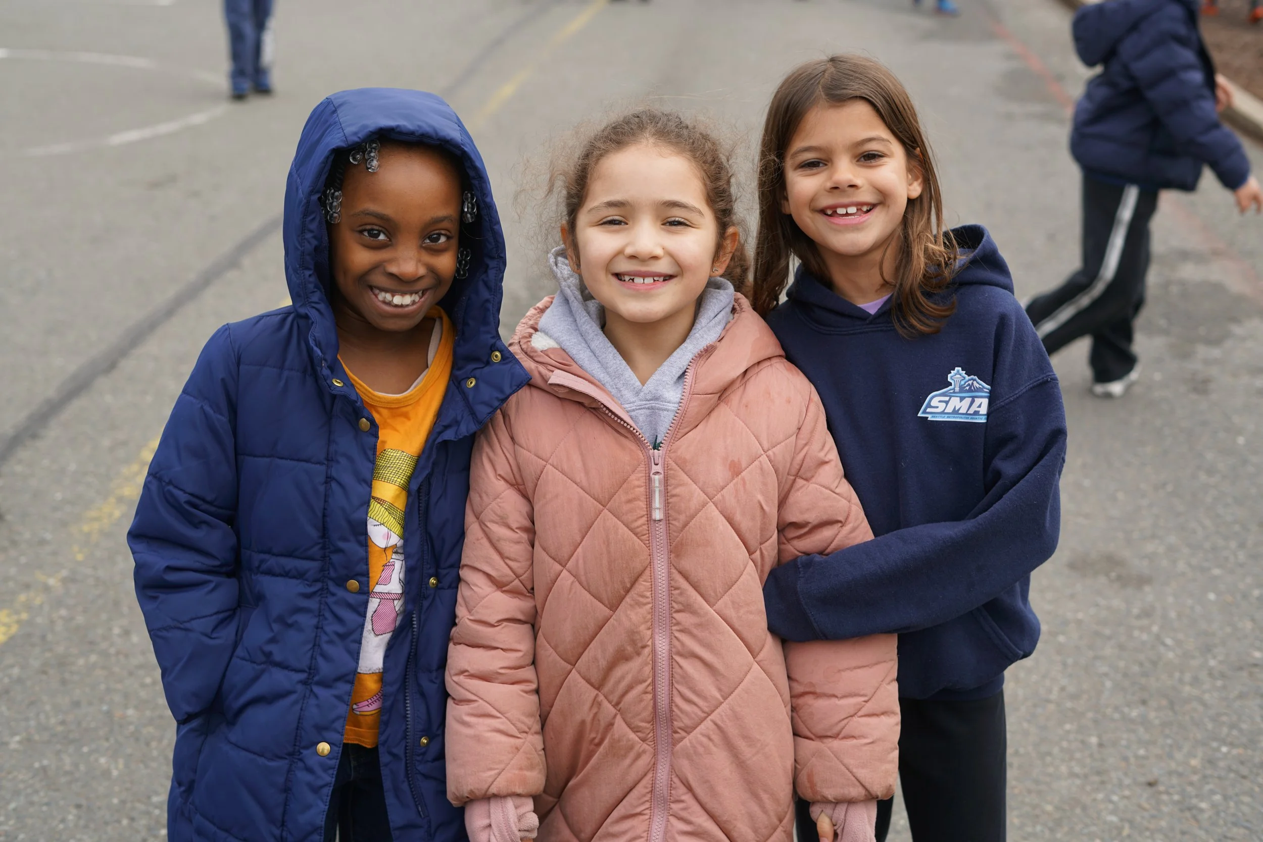 Three young girls smiling and standing close together outdoors, with two boys in jackets visible in the background on a paved area.