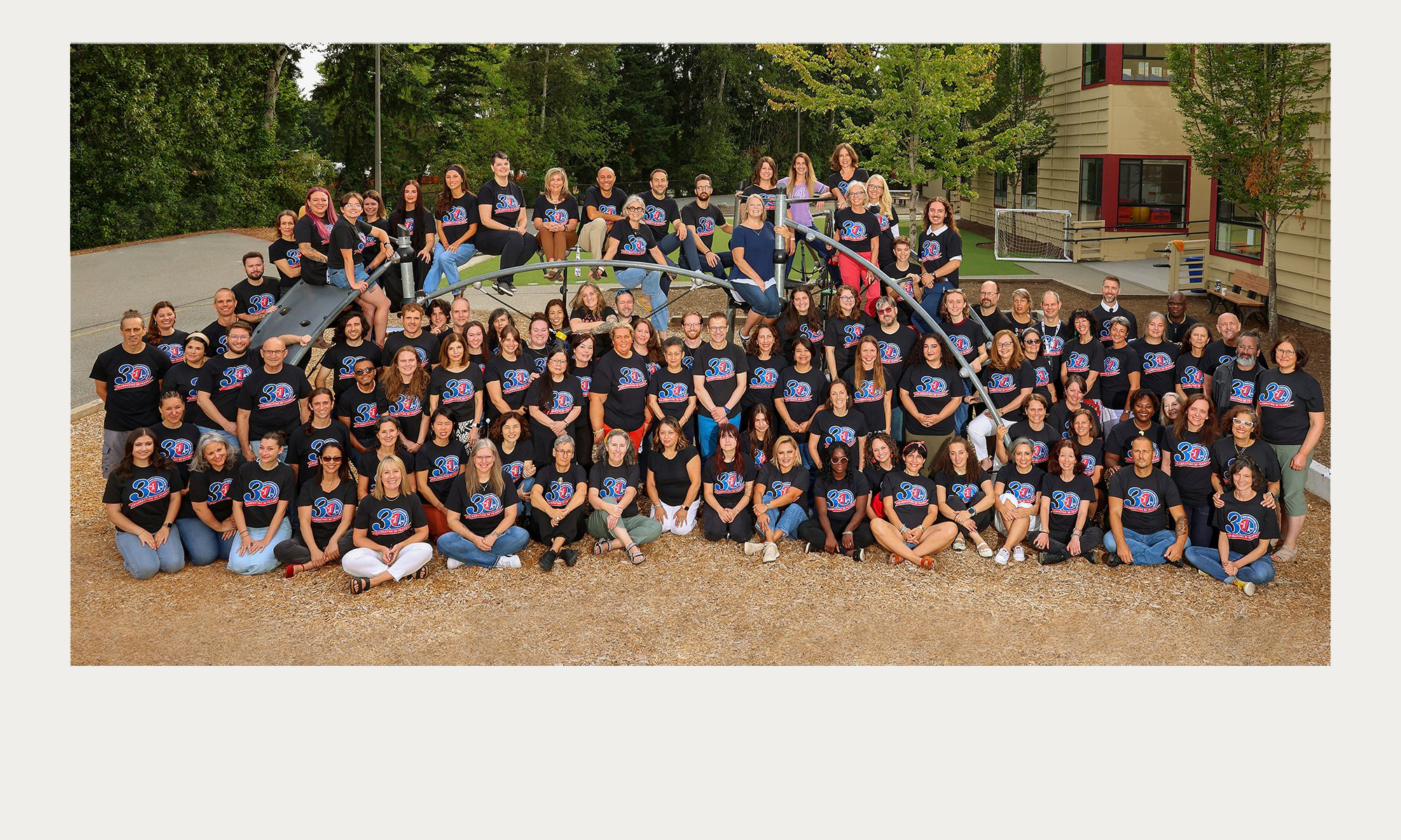Group of diverse people wearing black T-shirts with a 30 logo, gathered outdoors on a playground, some sitting on a slide, with trees and buildings in the background.