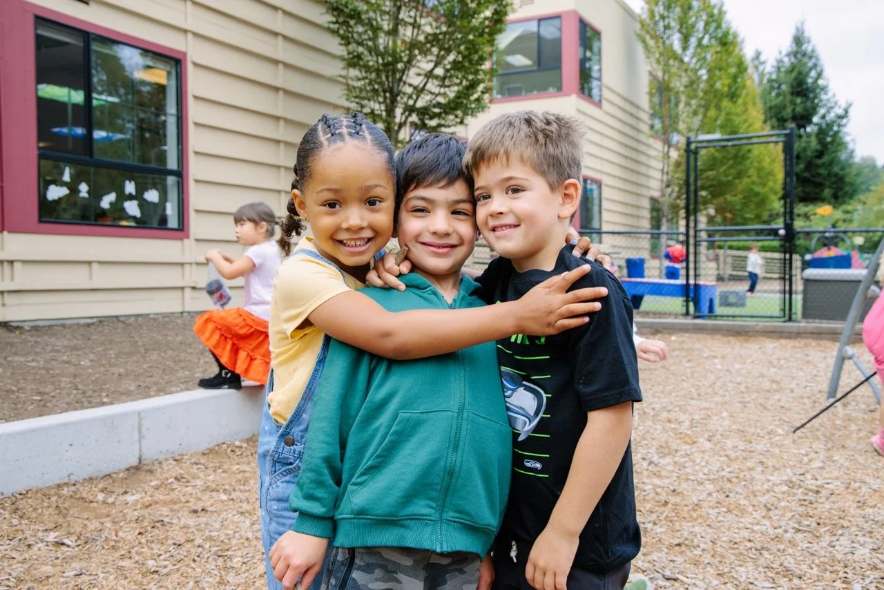 Three children smiling and hugging outdoors in a playground area, with other children playing in the background.