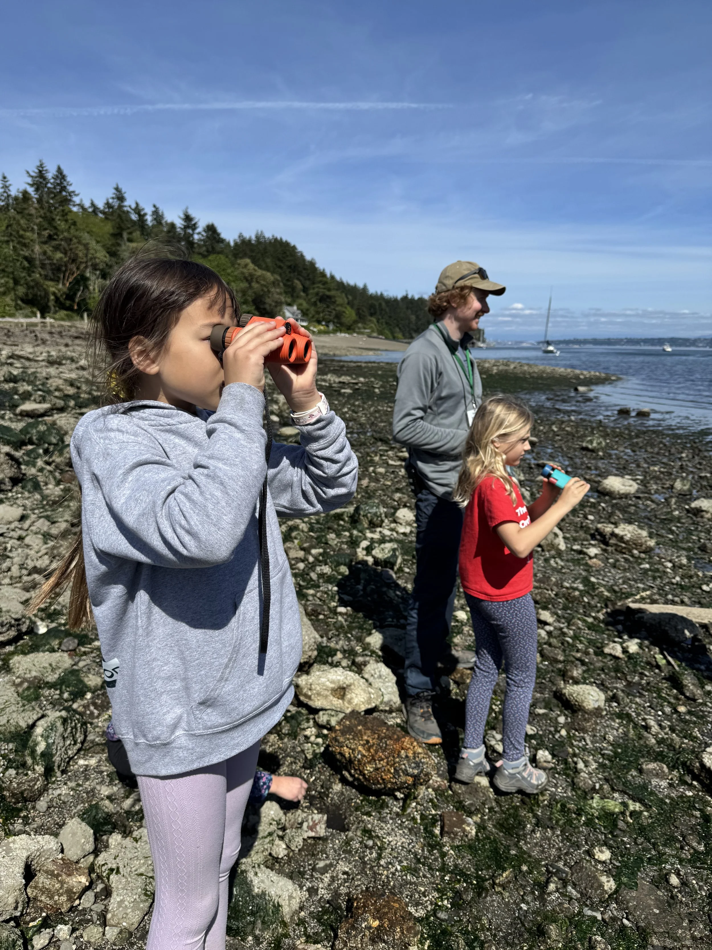 Children and a man standing on rocky shoreline along the water, looking through binoculars under a blue sky with some clouds.