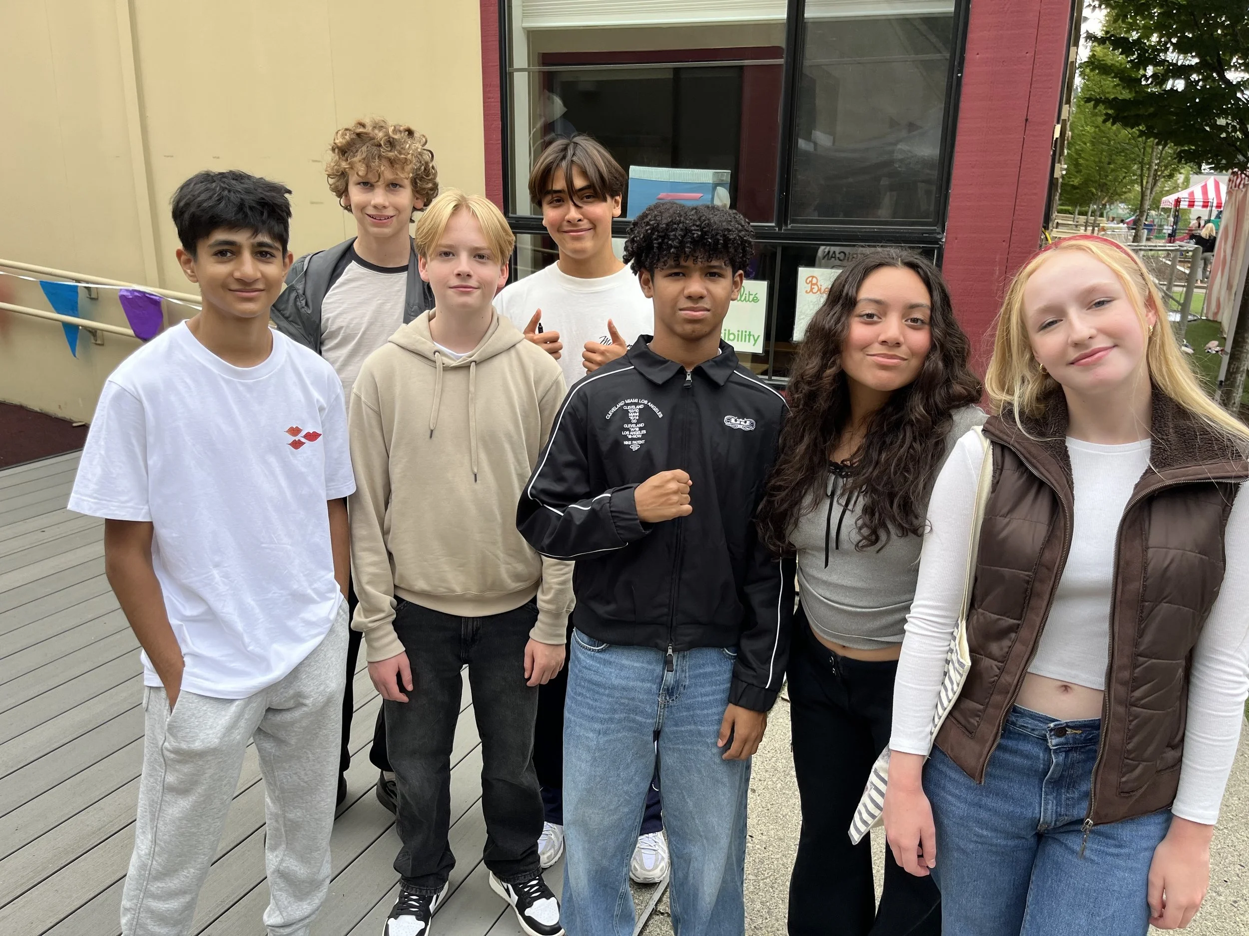 A group of nine diverse teenagers, six boys and three girls, standing on a wooden deck outdoors, smiling and posing for a photo. There are trees and a red building with large windows and a sign in the background.