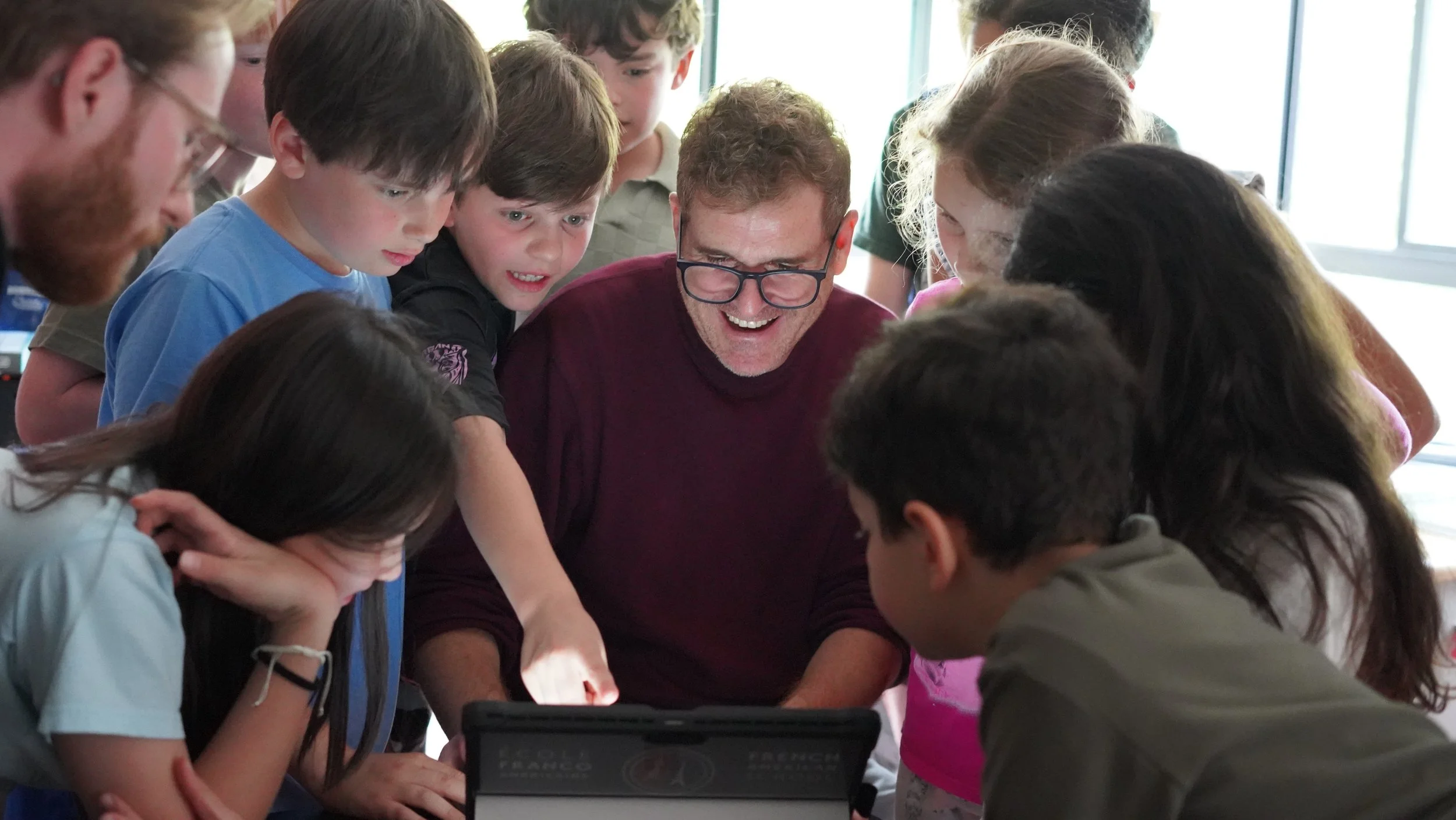 A group of children and one adult gathered around a table, closely examining an object or screen in a bright room with large windows.