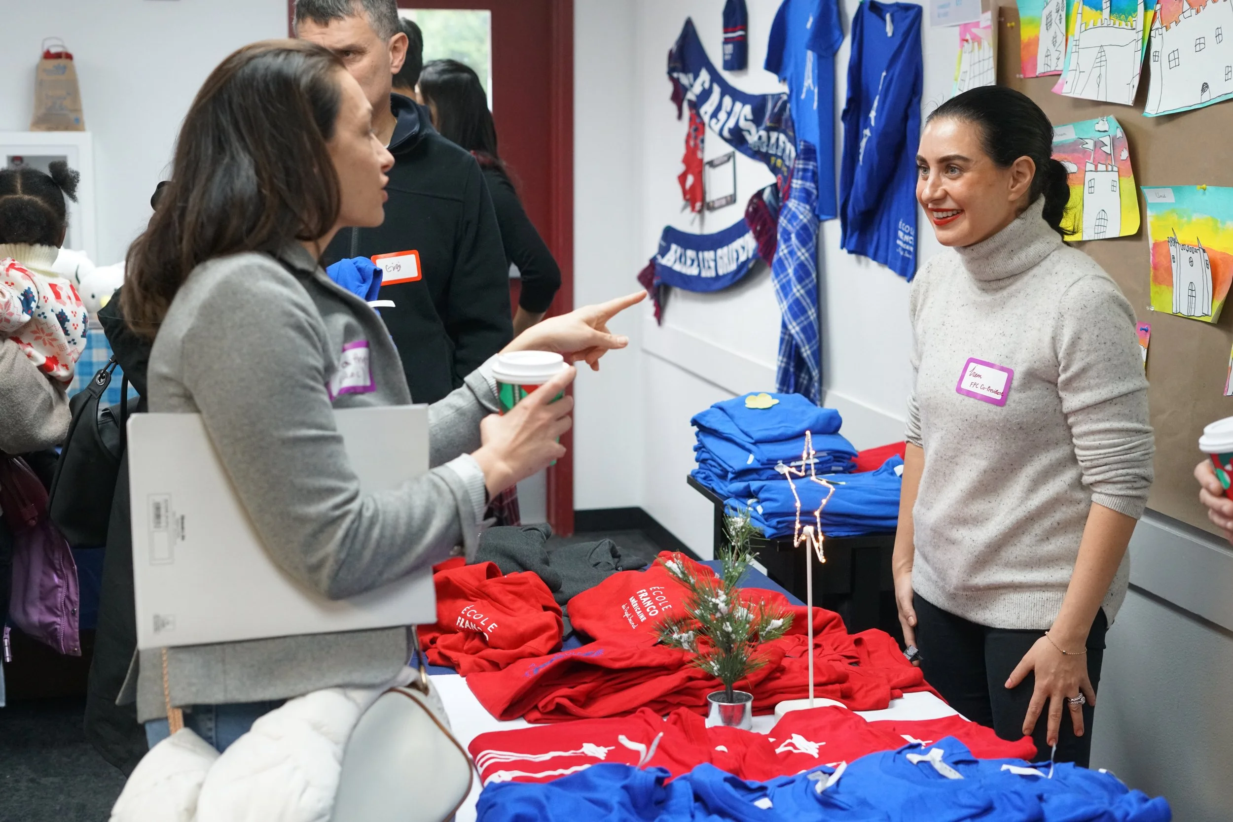Several people are at a booth with red and blue clothing items, some with white text, on a table. A woman with dark hair, wearing a beige turtleneck sweater and red lipstick, is smiling and talking to another woman holding a coffee cup and wearing a gray jacket. The background has colorful drawings and shirts hanging on the wall.