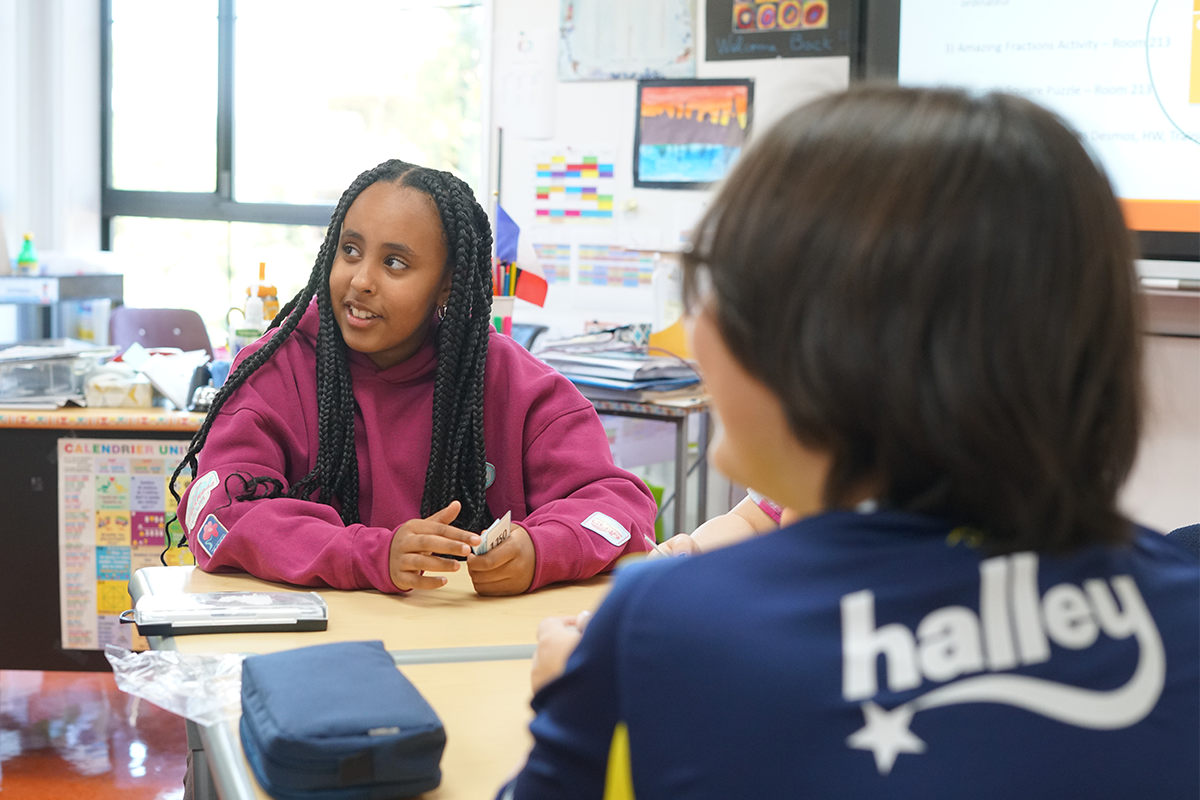 A young girl with braided hair wearing a pink hoodie sitting at a school desk, holding playing cards, and talking to a person with short hair wearing a navy shirt with 'haley' written on the back, in a colorful classroom.