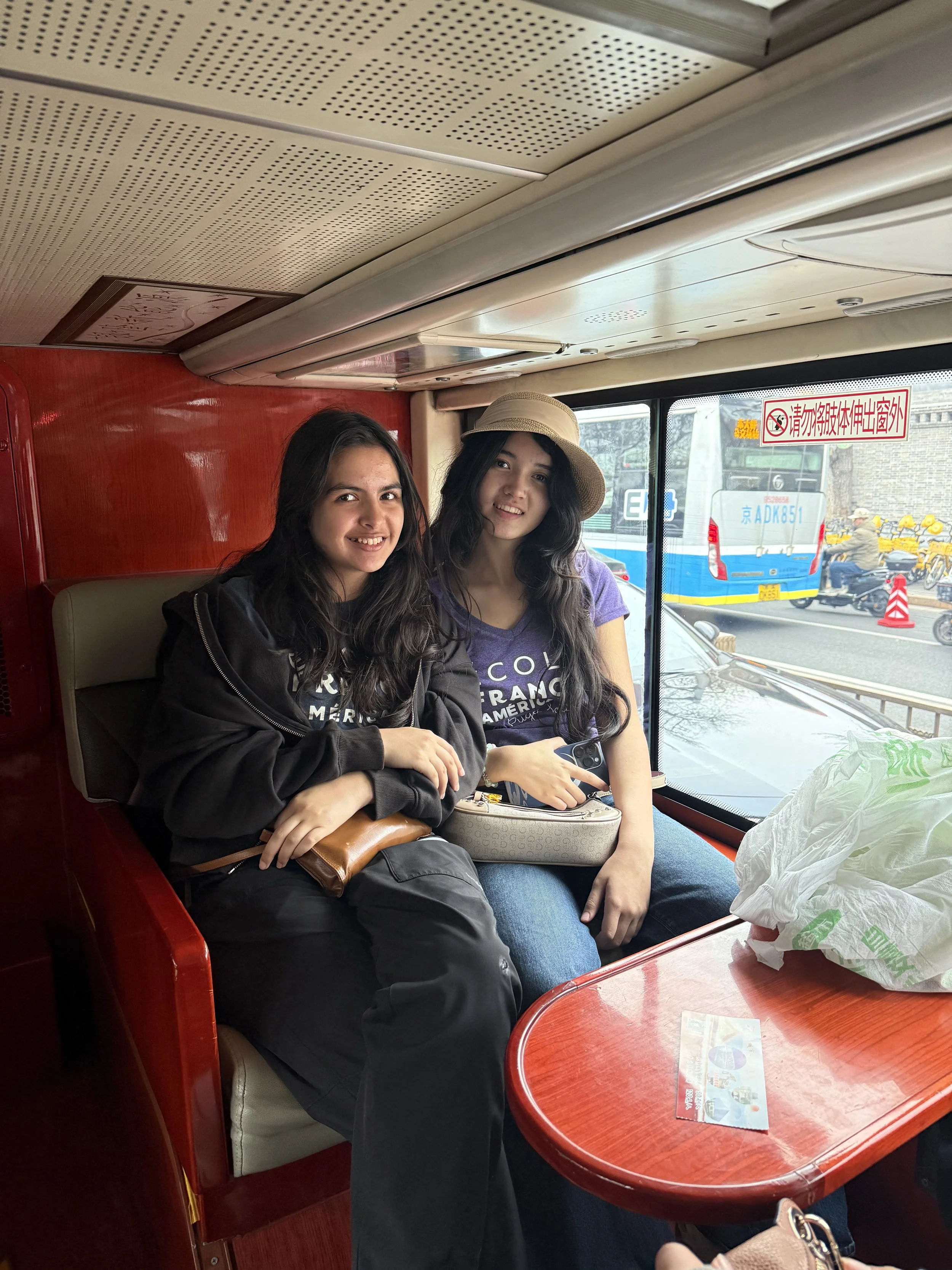 Two young women sitting close together on a bus, smiling at the camera. One has long dark hair and is wearing a black jacket, while the other has long dark hair and is wearing a purple shirt and a sun hat. A window behind them shows a busy street with a bus and motorcycles outside.