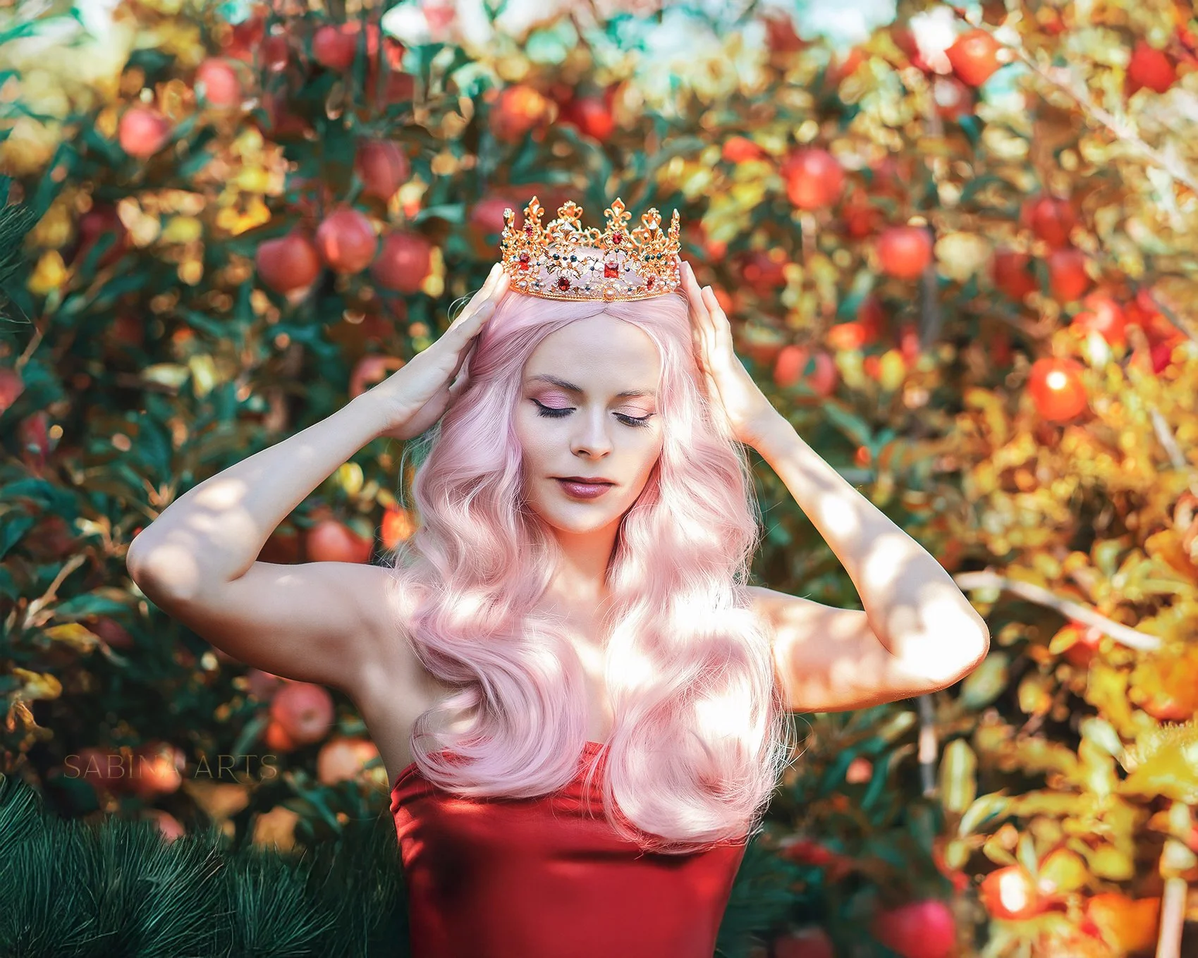 Woman with long pink hair wearing a golden crown with red and clear gemstones, standing in front of an apple orchard.