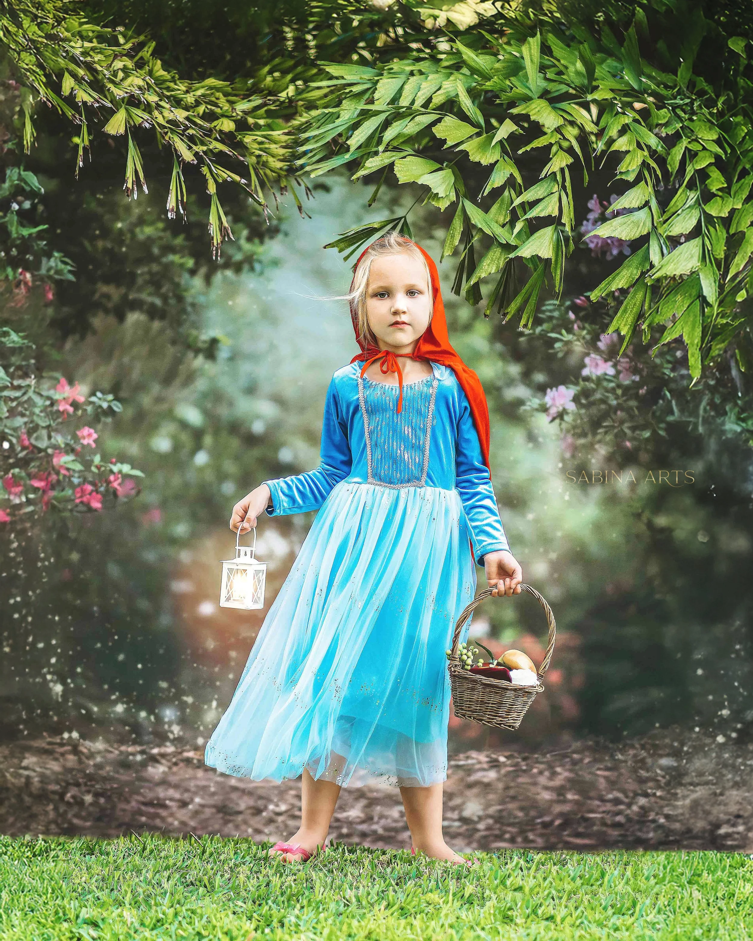 A young girl dressed as Little Red Riding Hood holding a lantern and a wicker basket outdoors among green leaves and pink flowers.