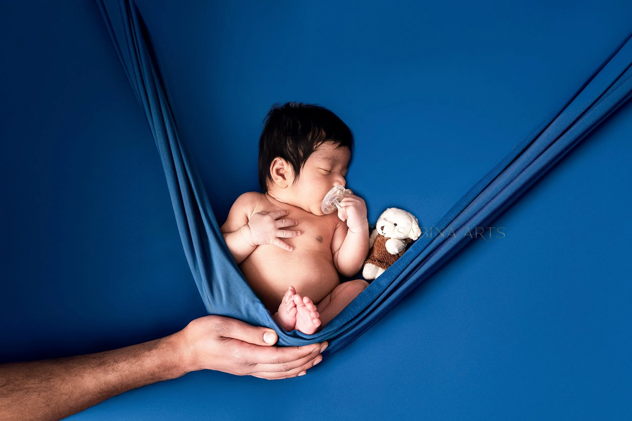 A newborn baby sleeping on a blue hammock, holding a pacifier and a small teddy bear, with an adult's hands gently supporting the hammock.