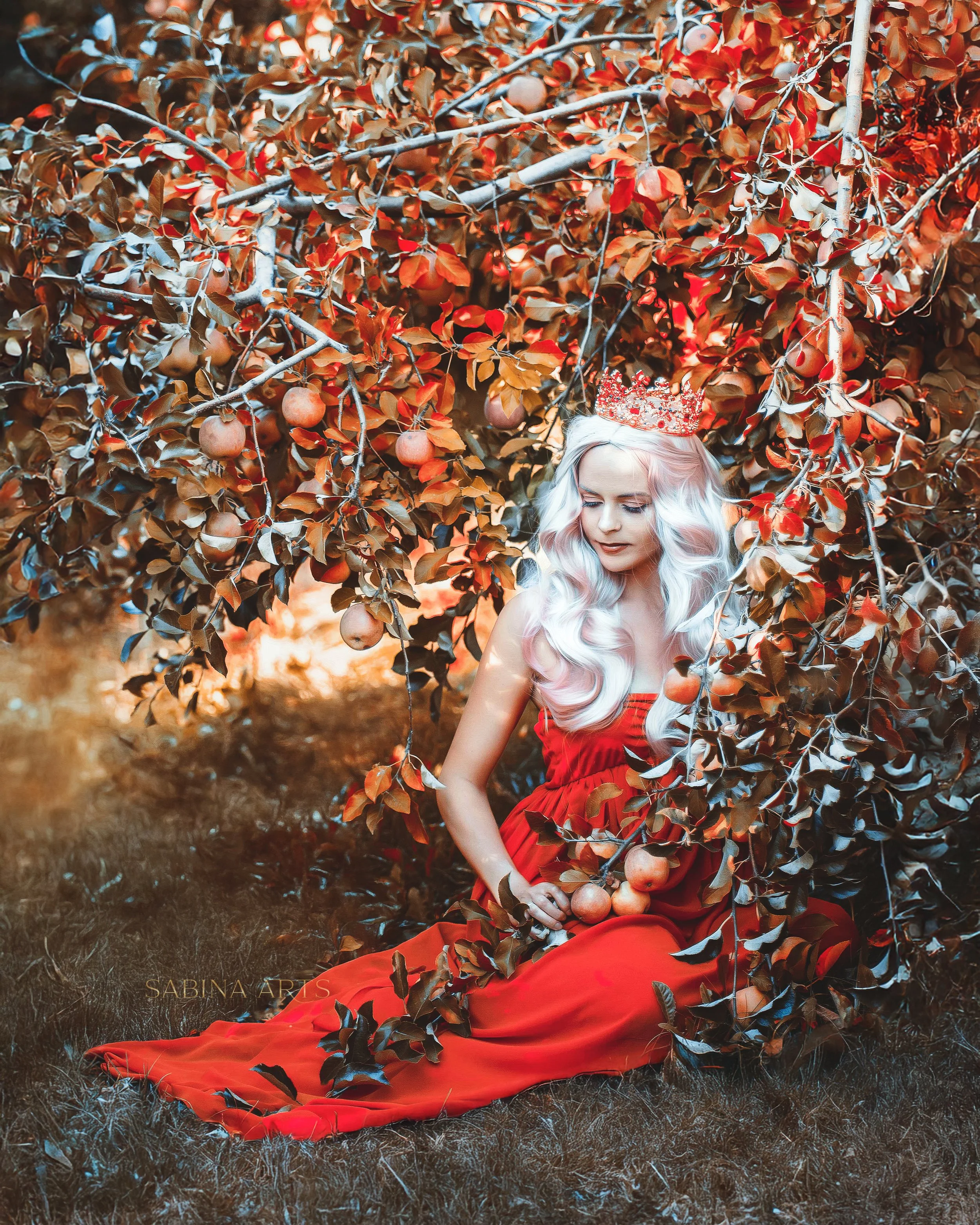 A woman with long, wavy, platinum blonde hair wearing a red dress and a sparkling tiara is sitting on the ground outdoors surrounded by a dense bush with autumn-colored leaves and small round apples. Her head is slightly tilted down with her eyes clo