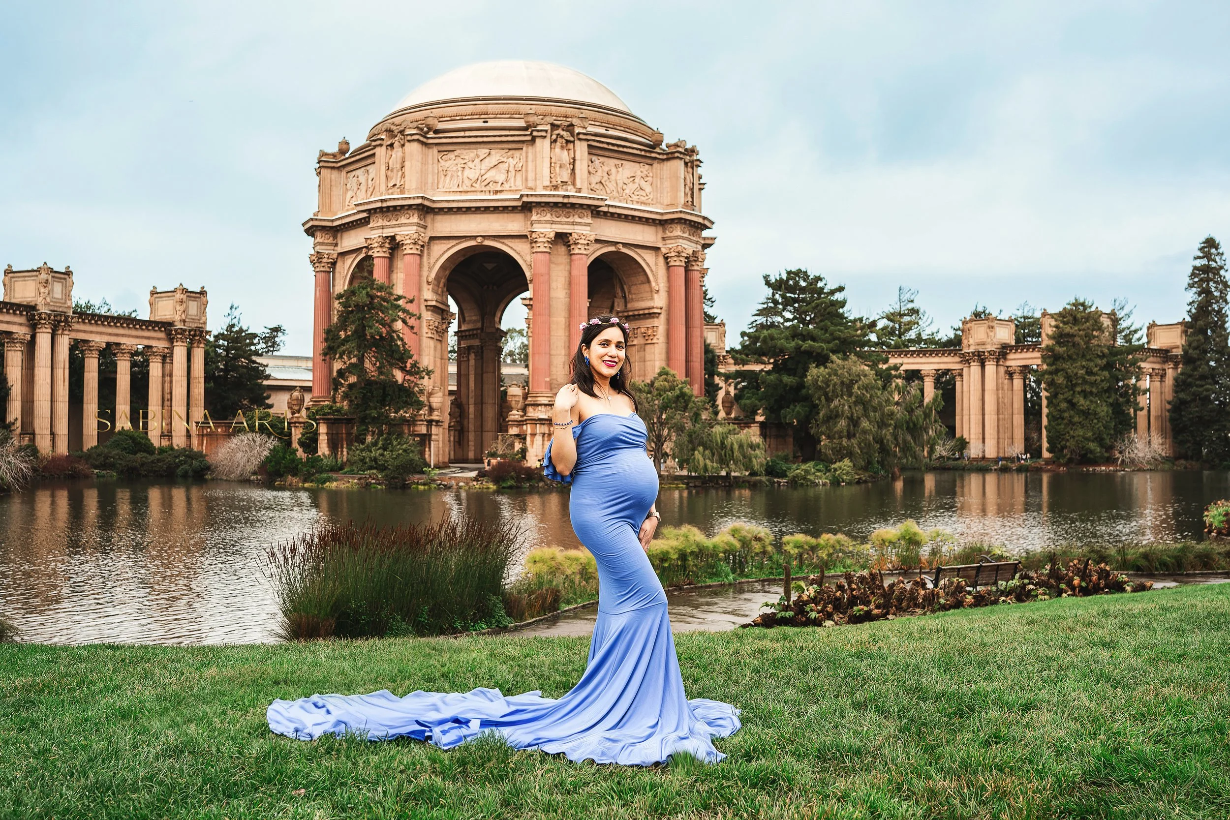 Pregnant woman in a blue gown standing on grass in front of a historic monument with columns and a dome, overlooking a water feature with trees and cloudy sky in the background.