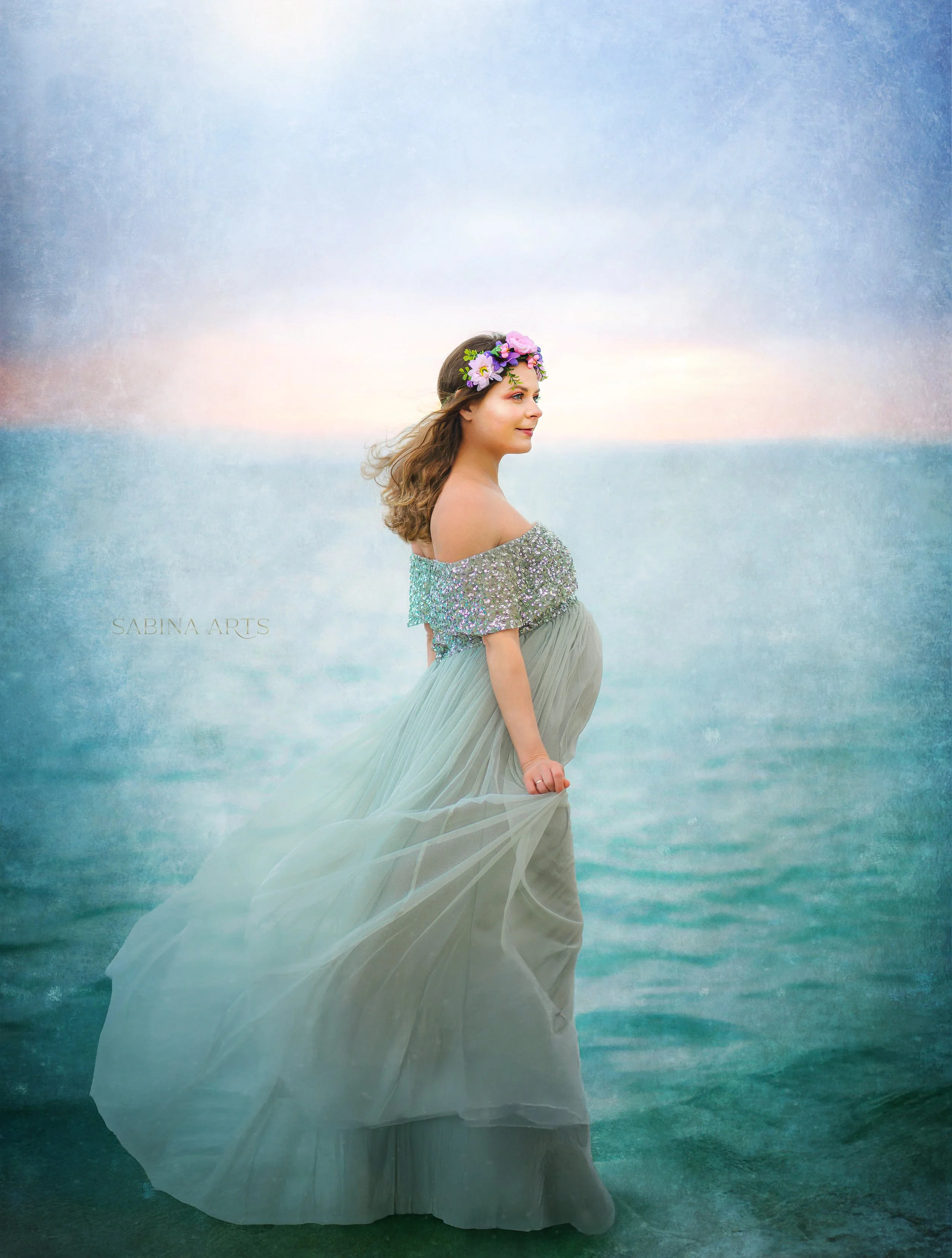 A woman with long hair wearing a flower crown and a bejeweled off-shoulder top stands in the ocean at sunset.