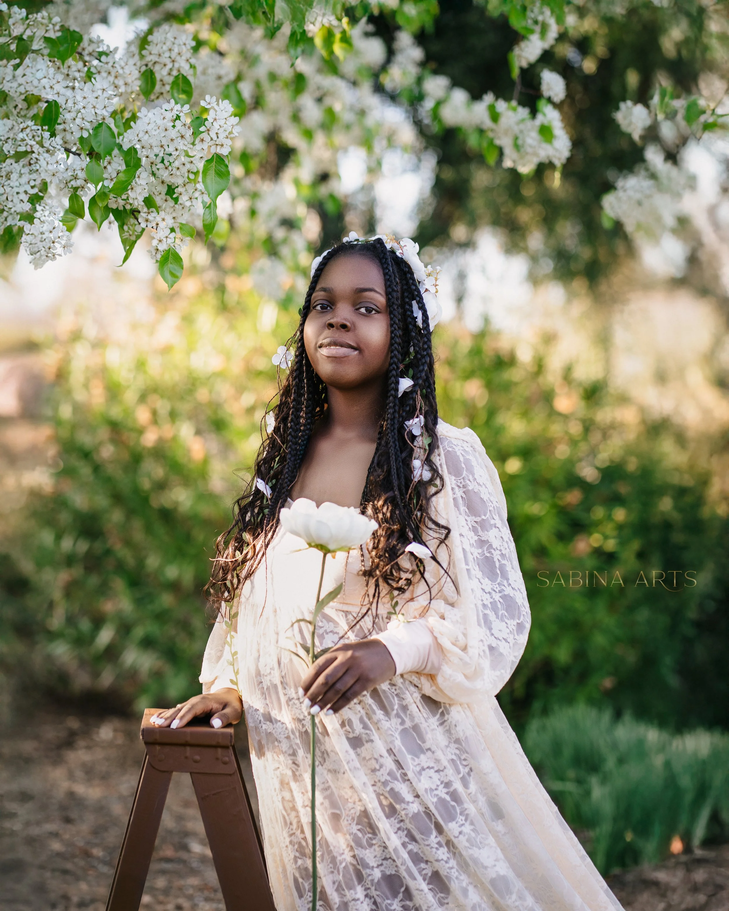 A young woman with braided hair adorned with white flowers, wearing a lace dress, holding a white flower, standing outdoors near blooming white flowers on trees in a natural setting.