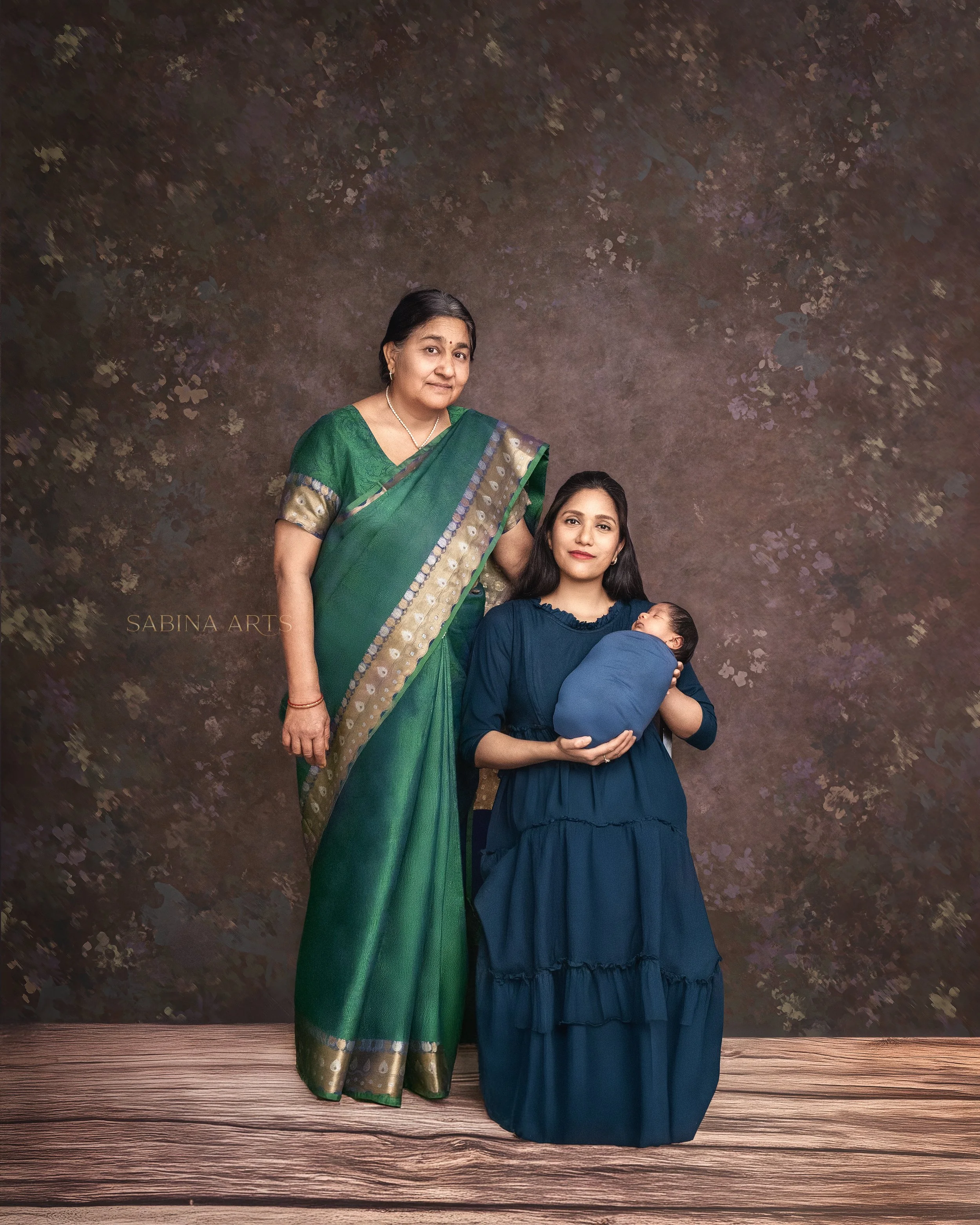 A woman in a green saree and a woman in a blue dress holding a baby, standing against a textured brown background with floral patterns.