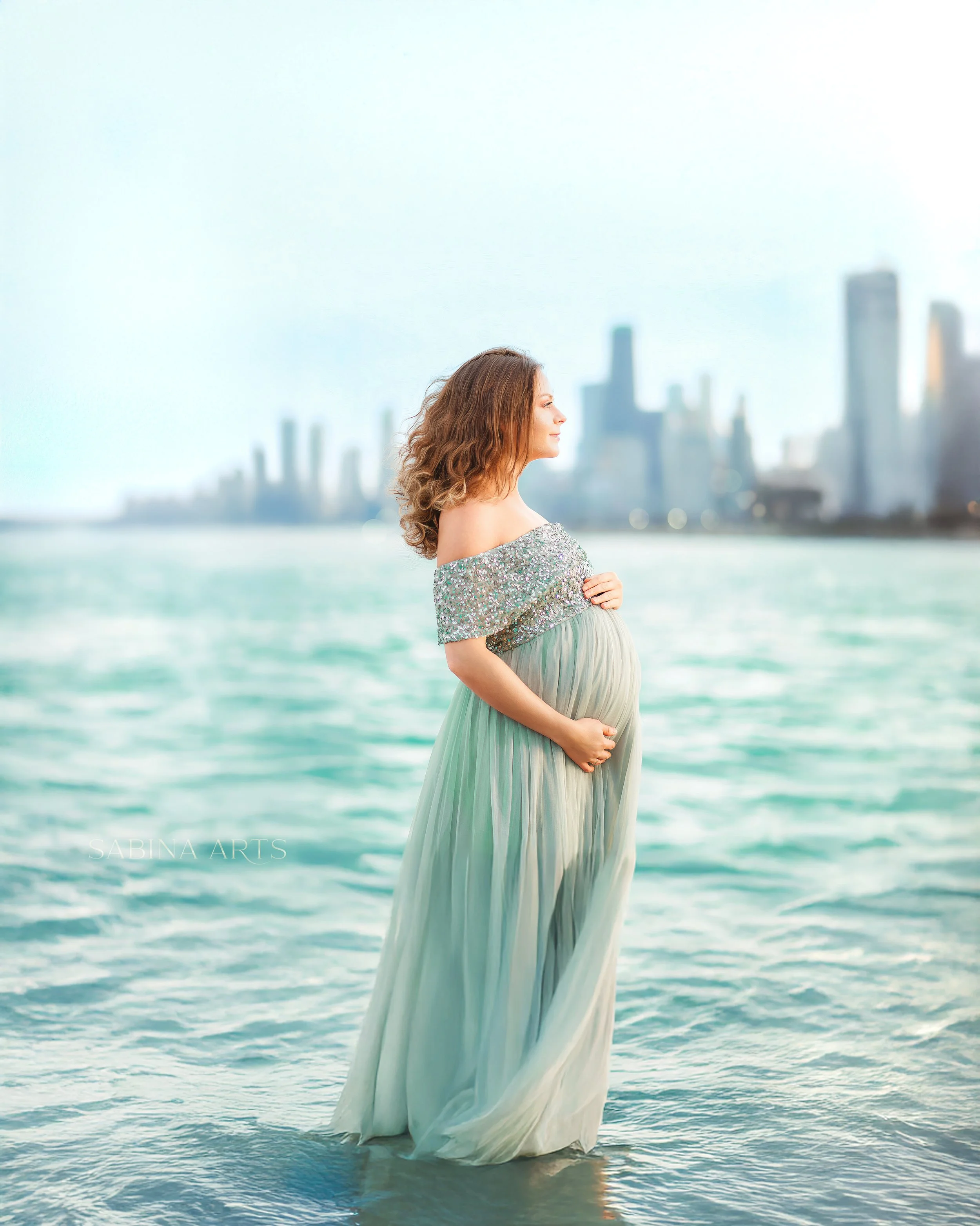 A pregnant woman with long wavy hair stands in shallow water at the beach, wearing a sparkling off-the-shoulder dress, with a city skyline in the background.
