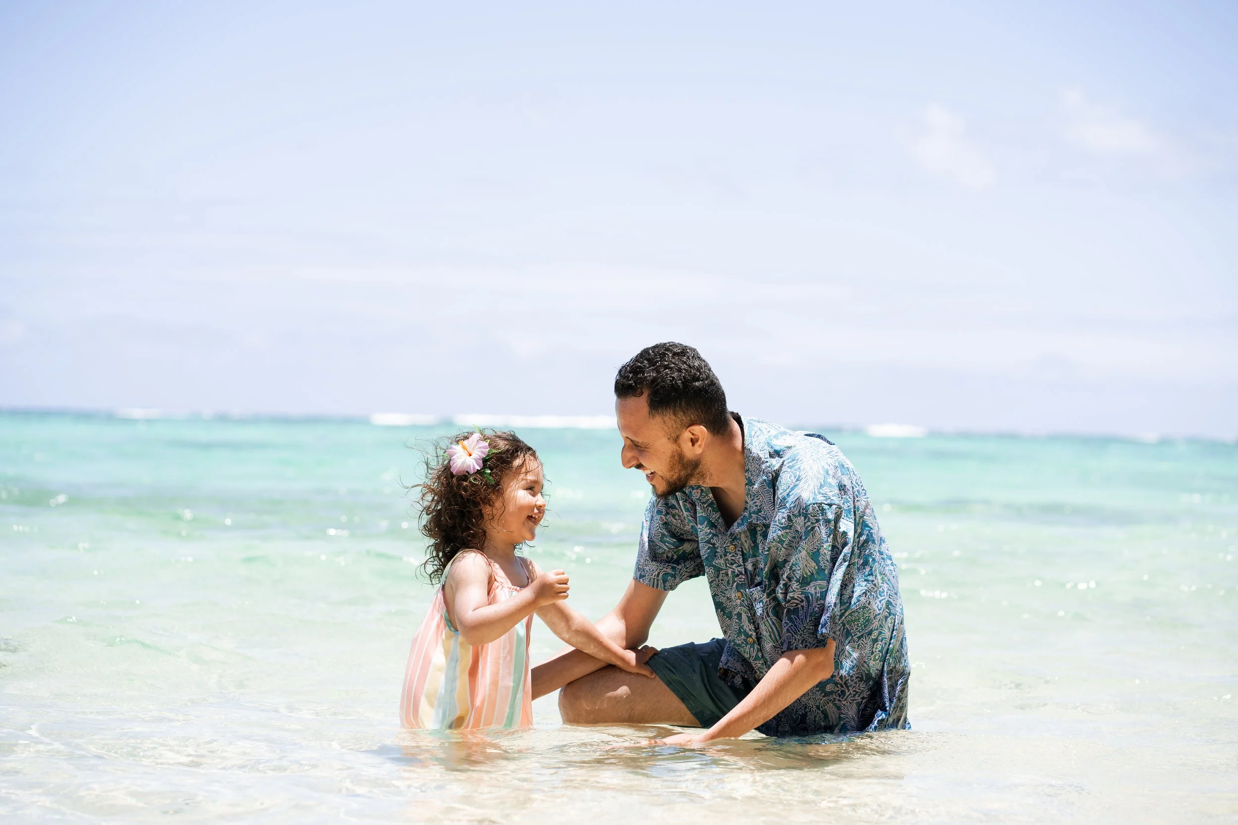 A man and a young girl enjoying time at the beach, smiling and holding hands in the shallow water with the ocean and sky in the background.