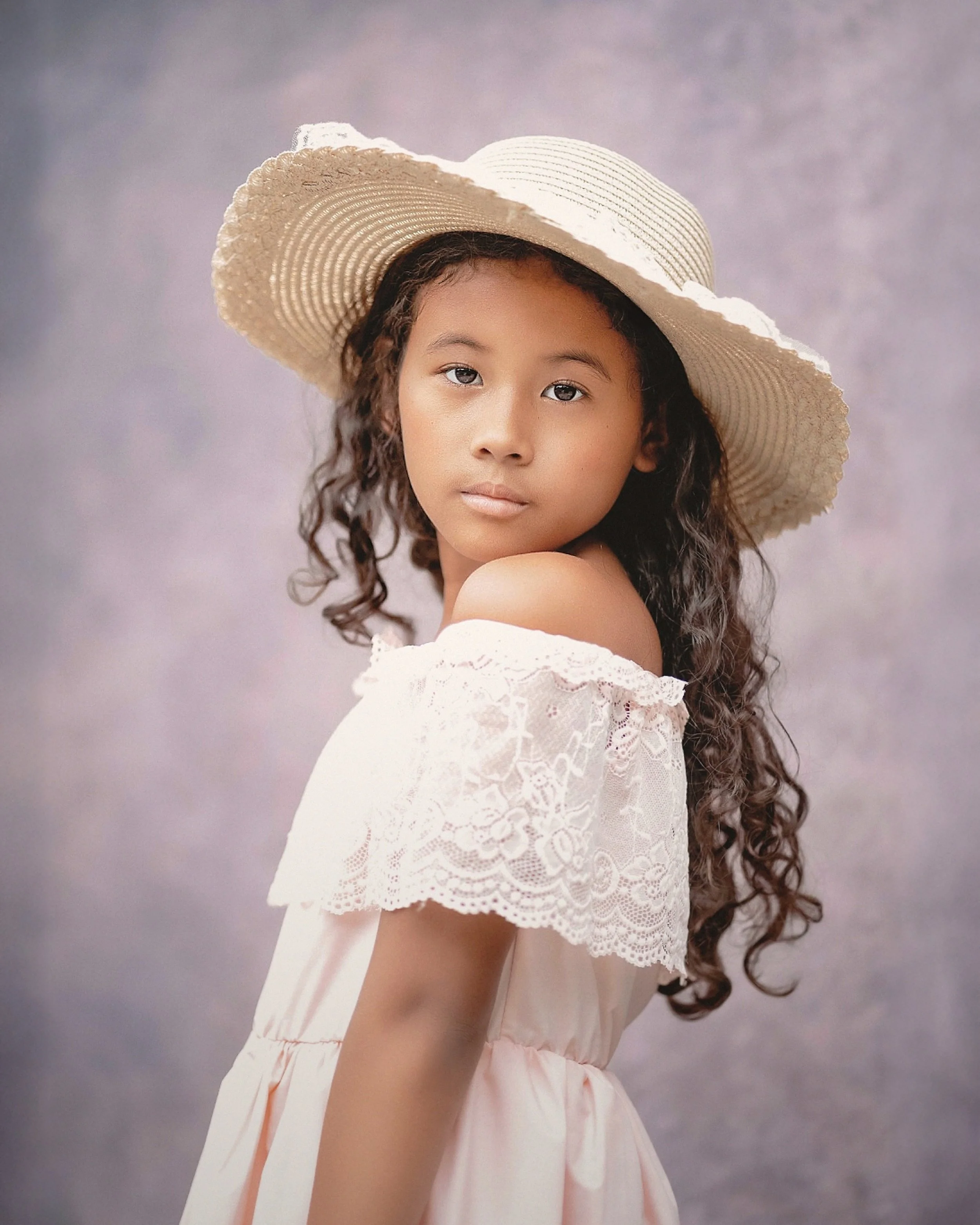 A young girl with long curly hair wearing a wide-brimmed straw hat and a white off-the-shoulder dress with lace details, standing against a cloudy sky background.