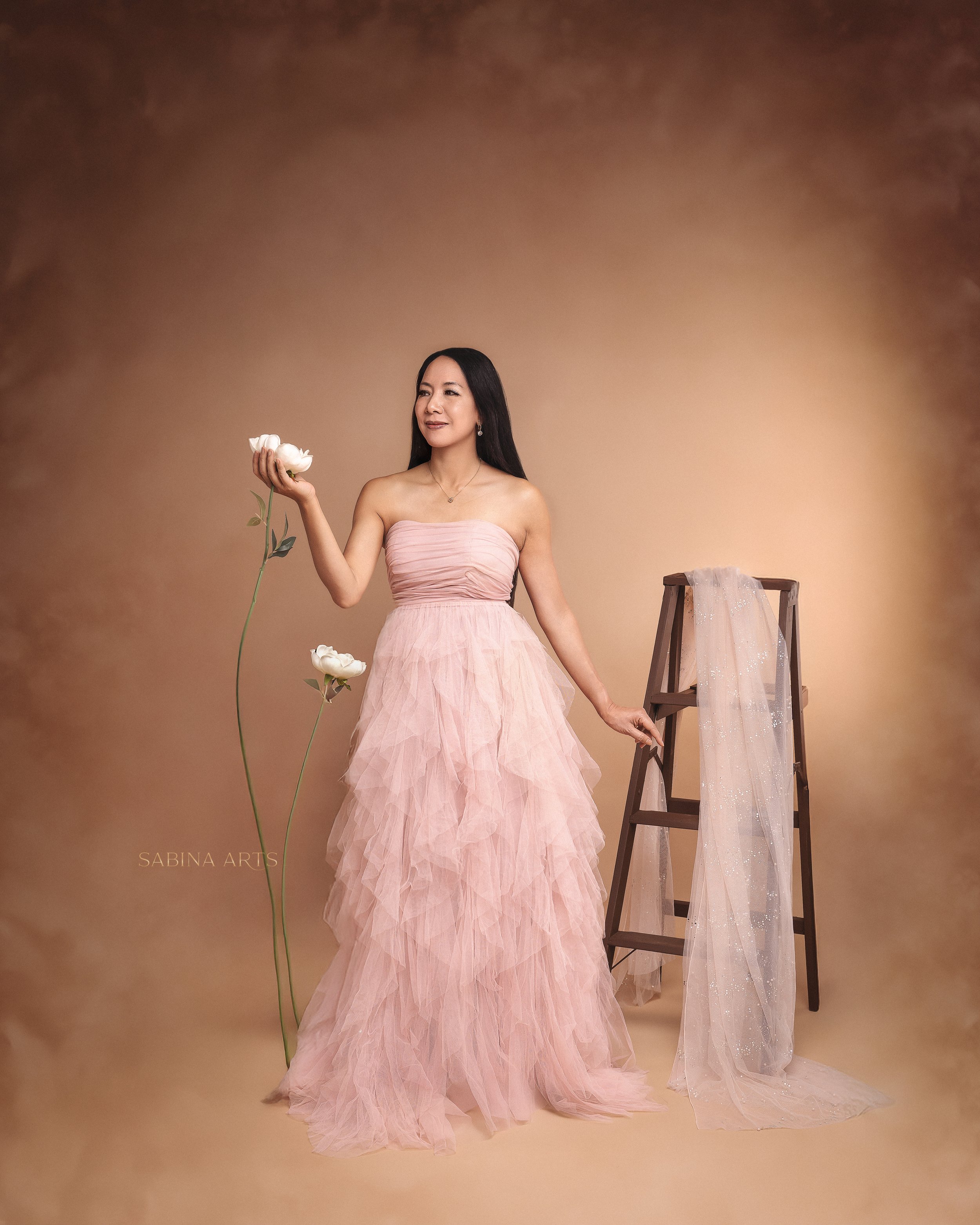 A woman in a strapless pink gown holding white flowers, standing in front of a plain beige background with a wooden ladder draped with a sheer fabric on the right side.