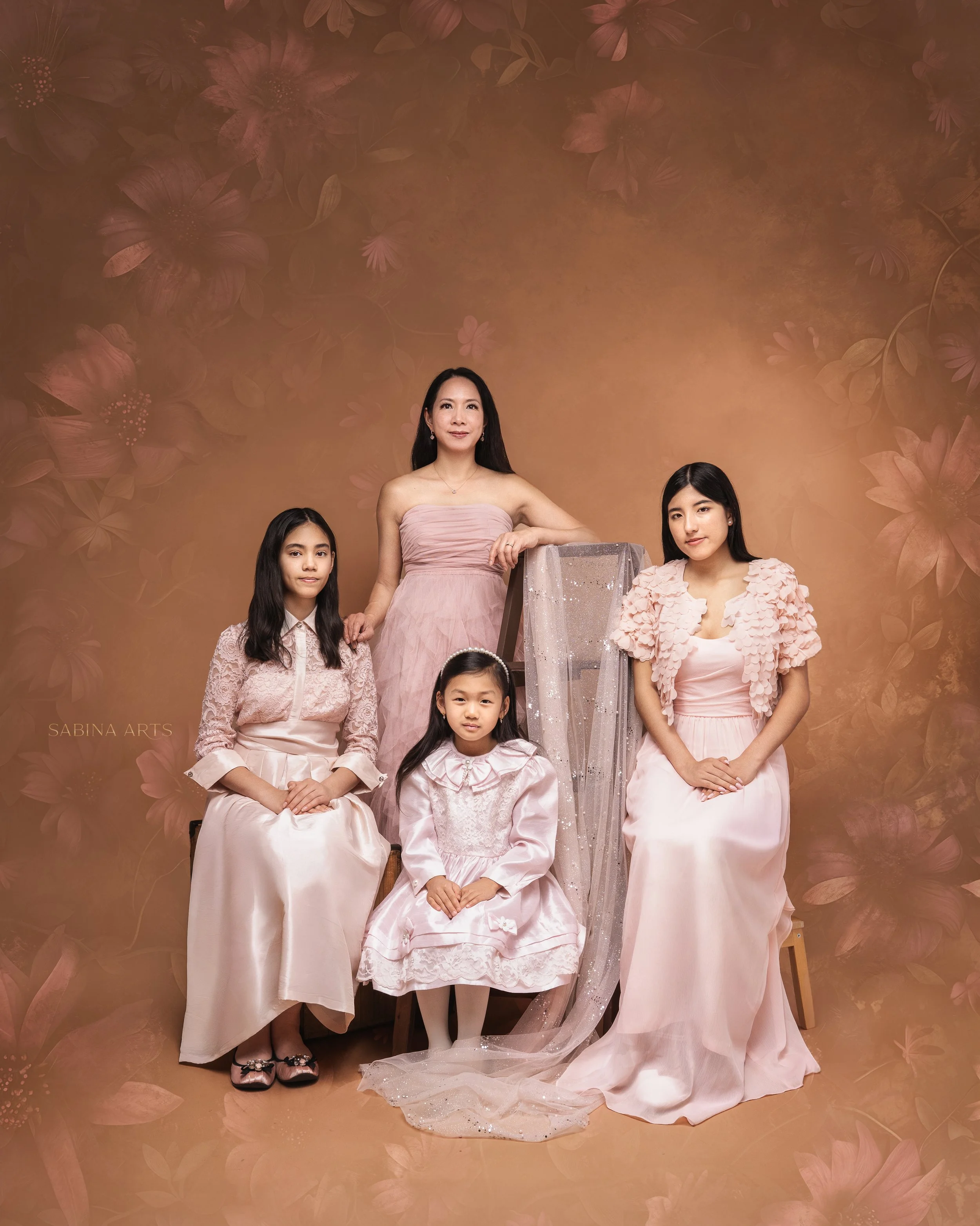 Group portrait of five females, including a woman and four girls, dressed in elegant pastel pink and beige dresses, standing against a floral patterned background.