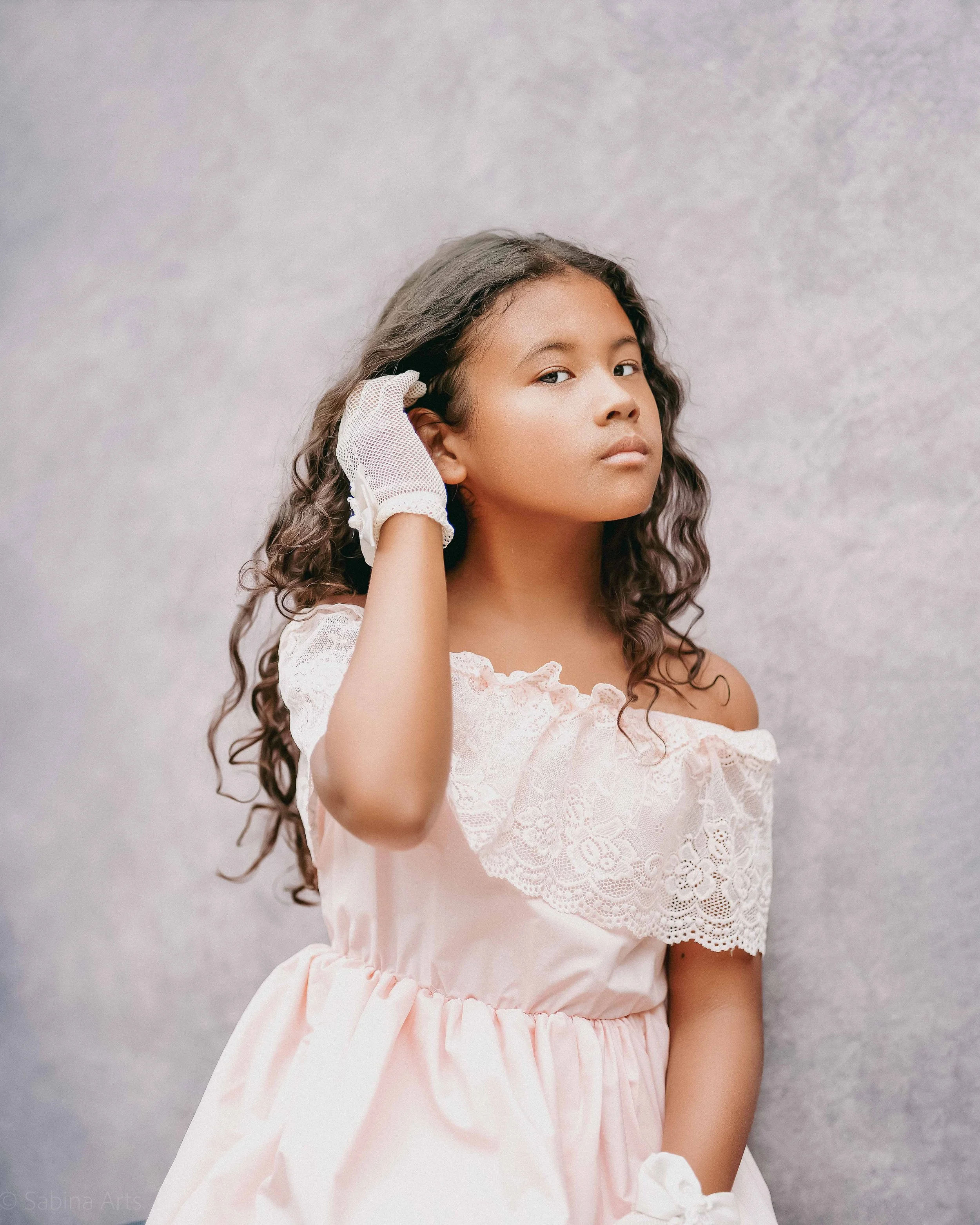 A girl with curly hair wearing a white lace dress and a white glove, standing against a grey textured background.