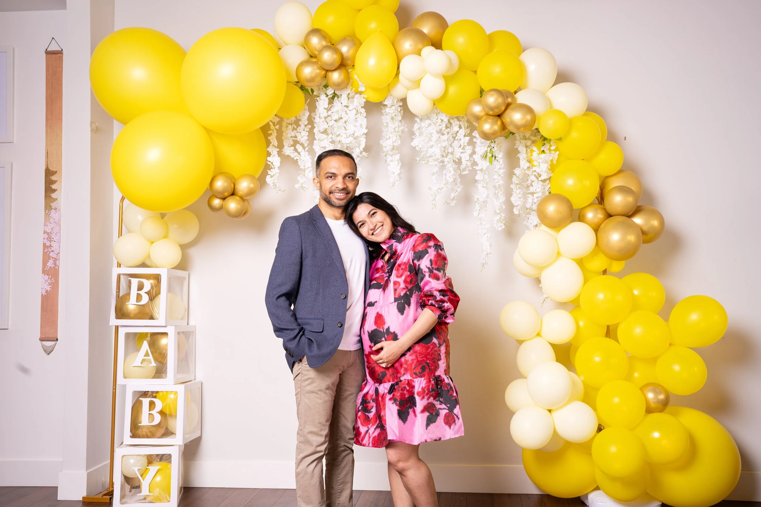 A smiling man and woman stand close together in front of a yellow balloon arch and a white decorative letter display spelling out 'BABY' with balloons inside the letters. The woman wears a pink floral dress, and the man wears a gray blazer and beige pants, celebrating a baby shower.