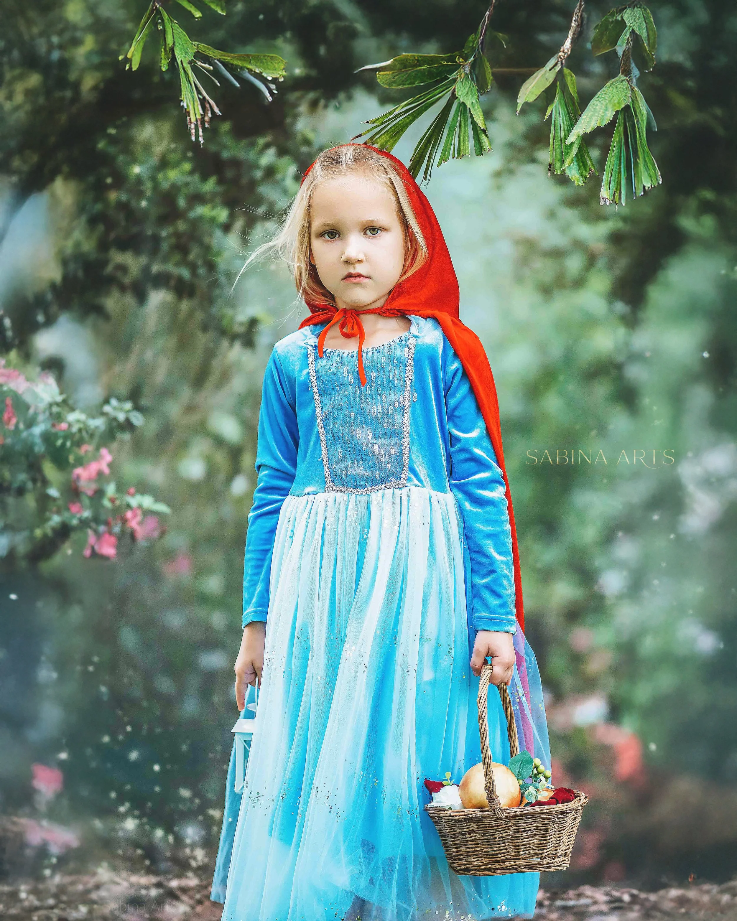 A young girl dressed as Little Red Riding Hood stands outdoors holding a basket containing bread and grapes, with a forest in the background.