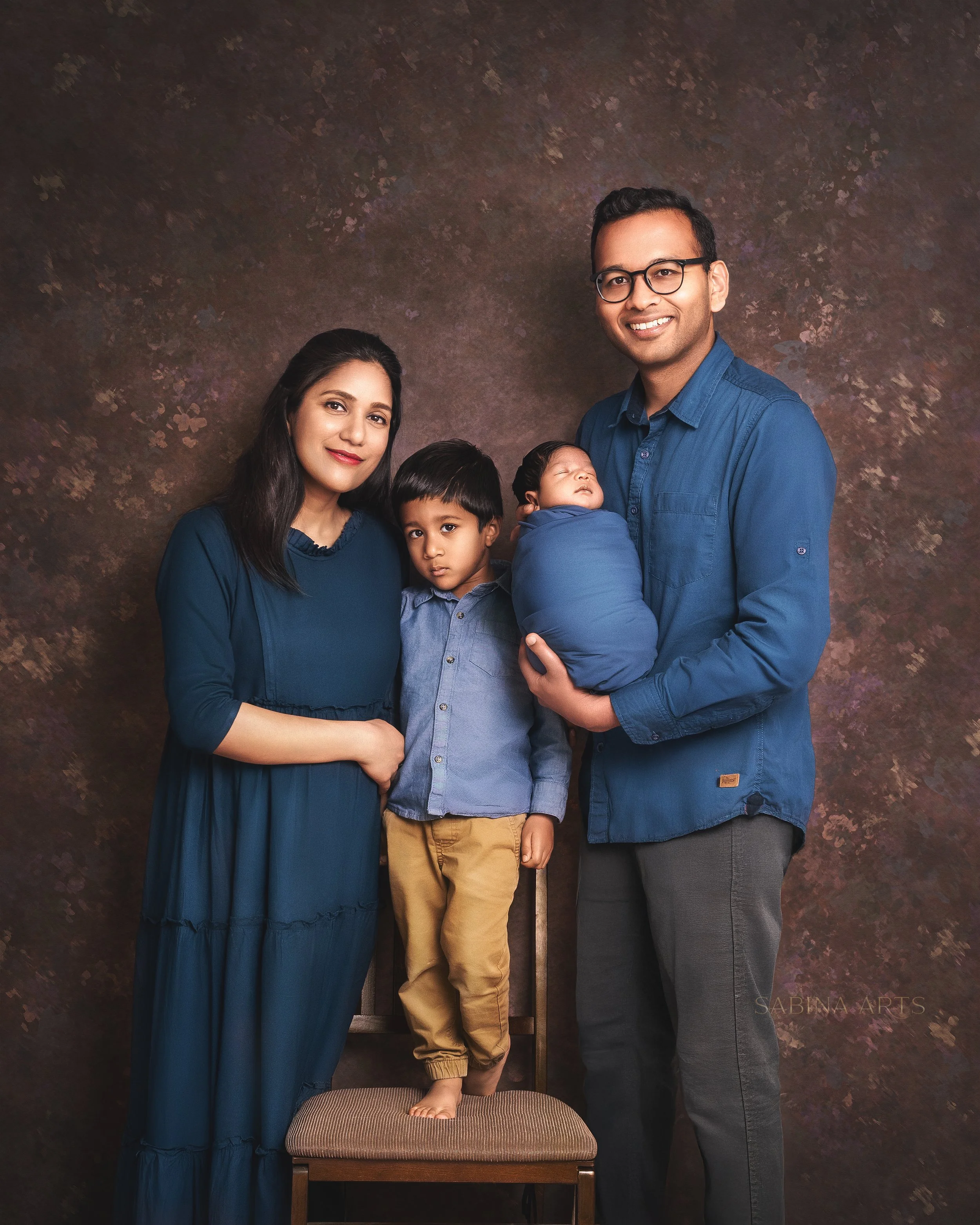 A family portrait of four people, including a woman, a man, a young boy, and a newborn baby, all dressed in blue, standing against a textured brown background. The woman has long dark hair, and the man has short dark hair and glasses. The boy and the