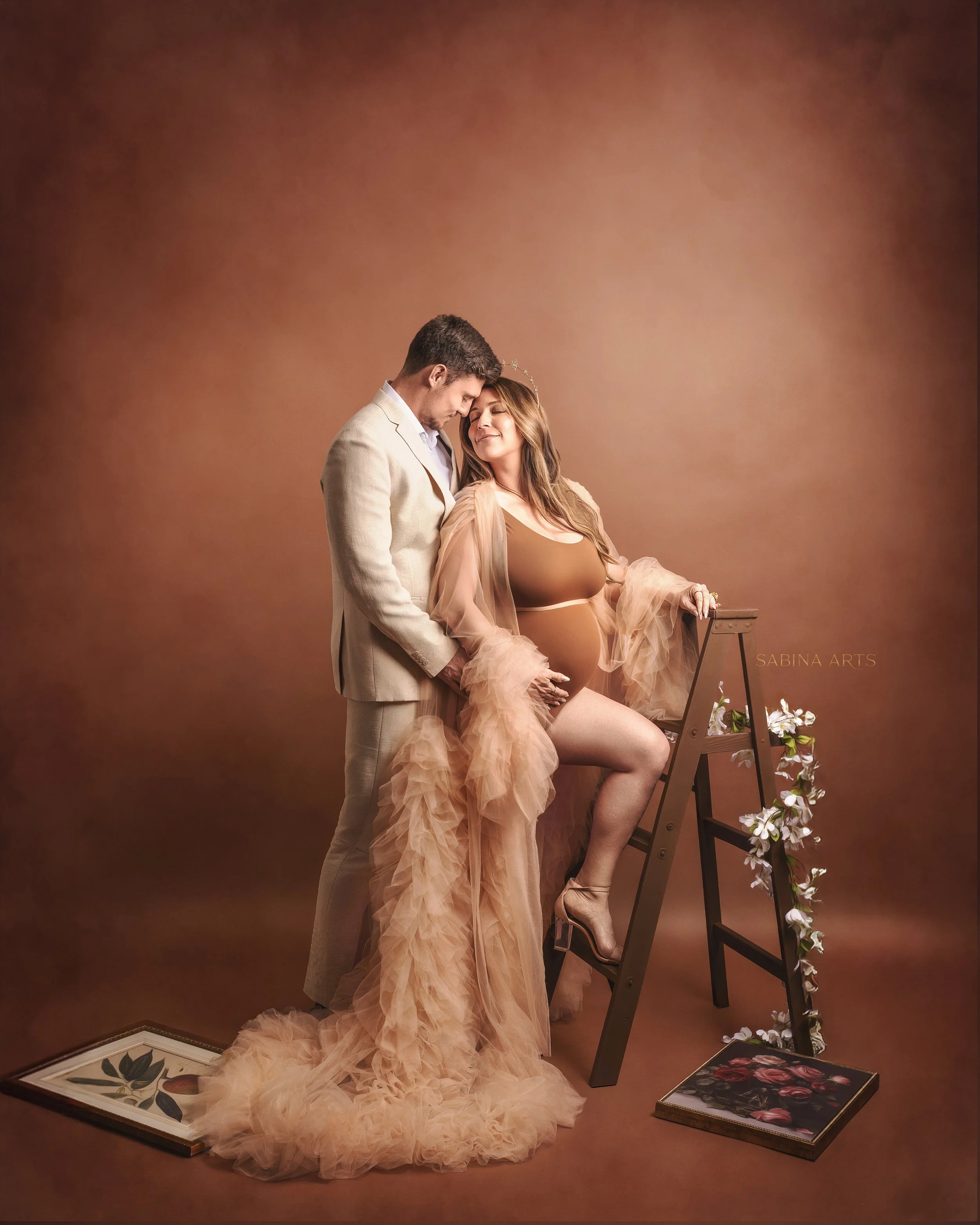 A man and a pregnant woman stand close together, with foreheads touching, in a warm-toned studio photograph. The woman wears a peach-colored sheer gown with ruffled fabric, and a tiara, while the man wears a light-colored suit. The woman is leaning o