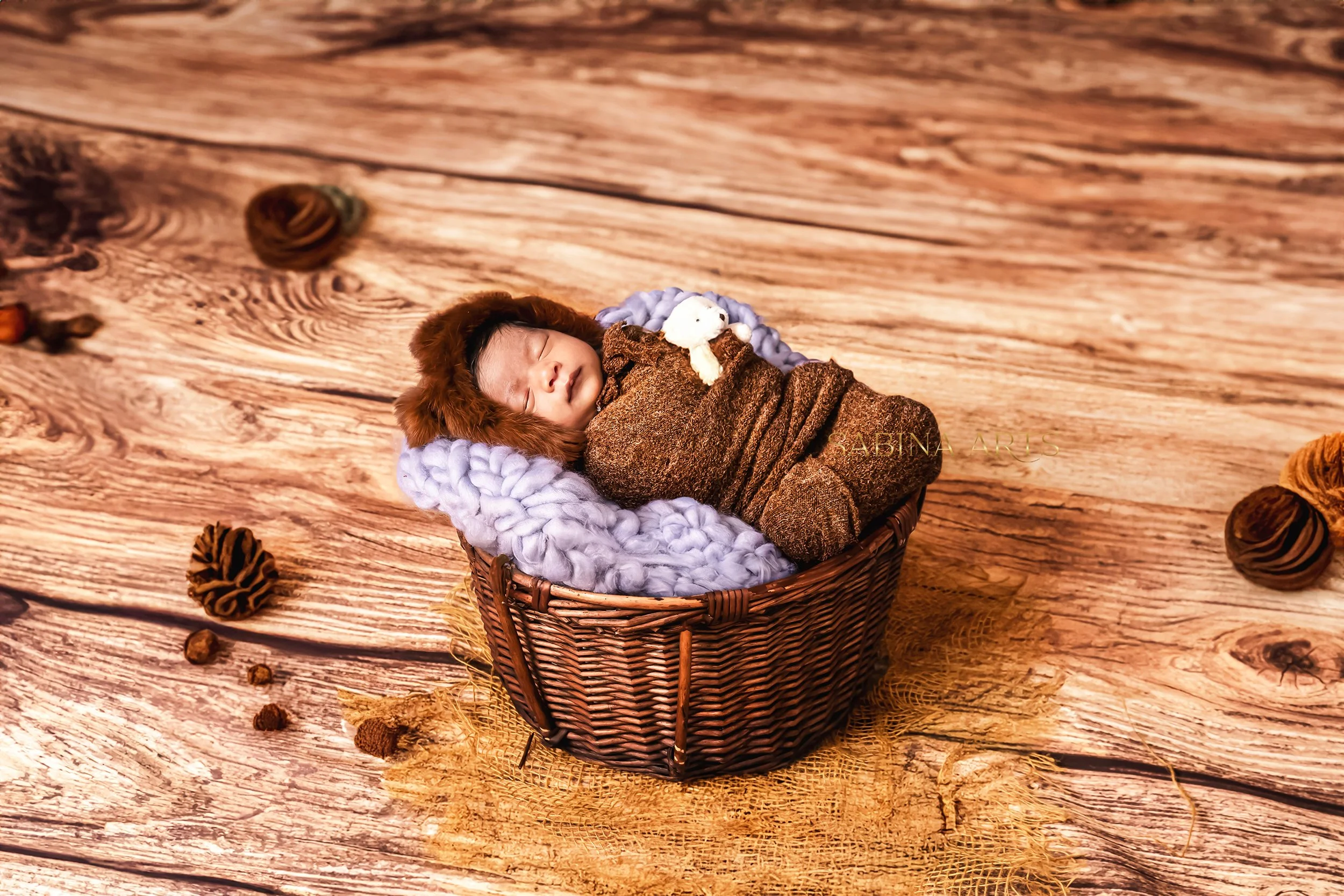 A sleeping baby dressed in a brown outfit with a bear hood, lying in a wicker basket lined with soft blankets, on a wooden floor with scattered pinecones around.