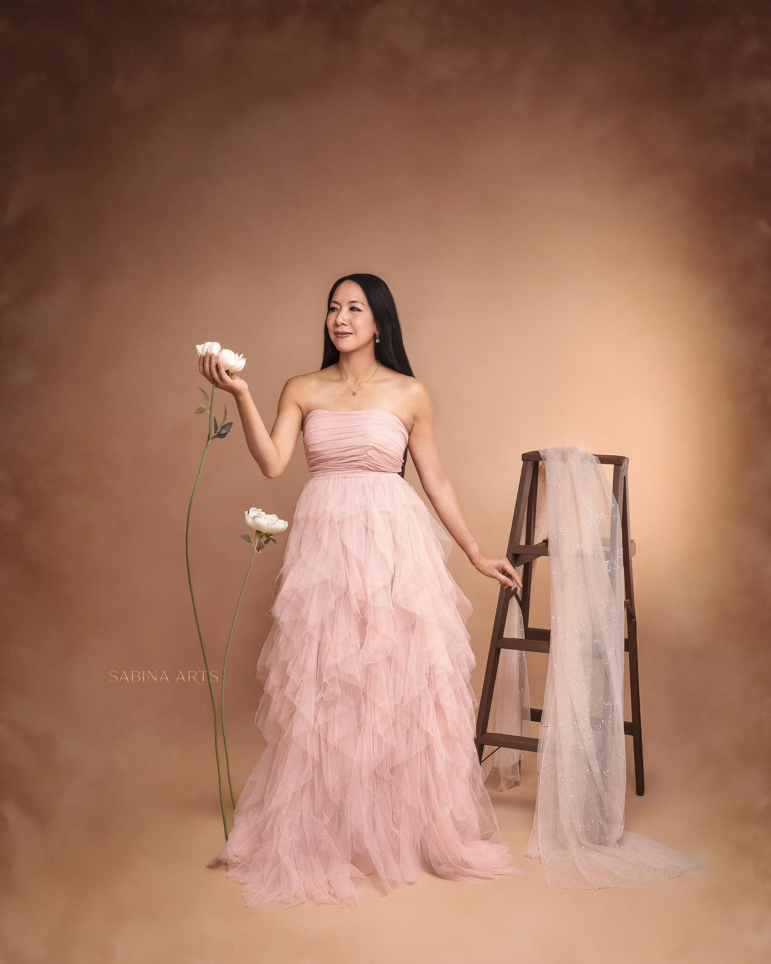 A woman in a strapless pink, tulle evening gown, holding a white flower in her right hand, standing against a beige background with a wooden ladder draped with a sheer, glittery fabric to her right.