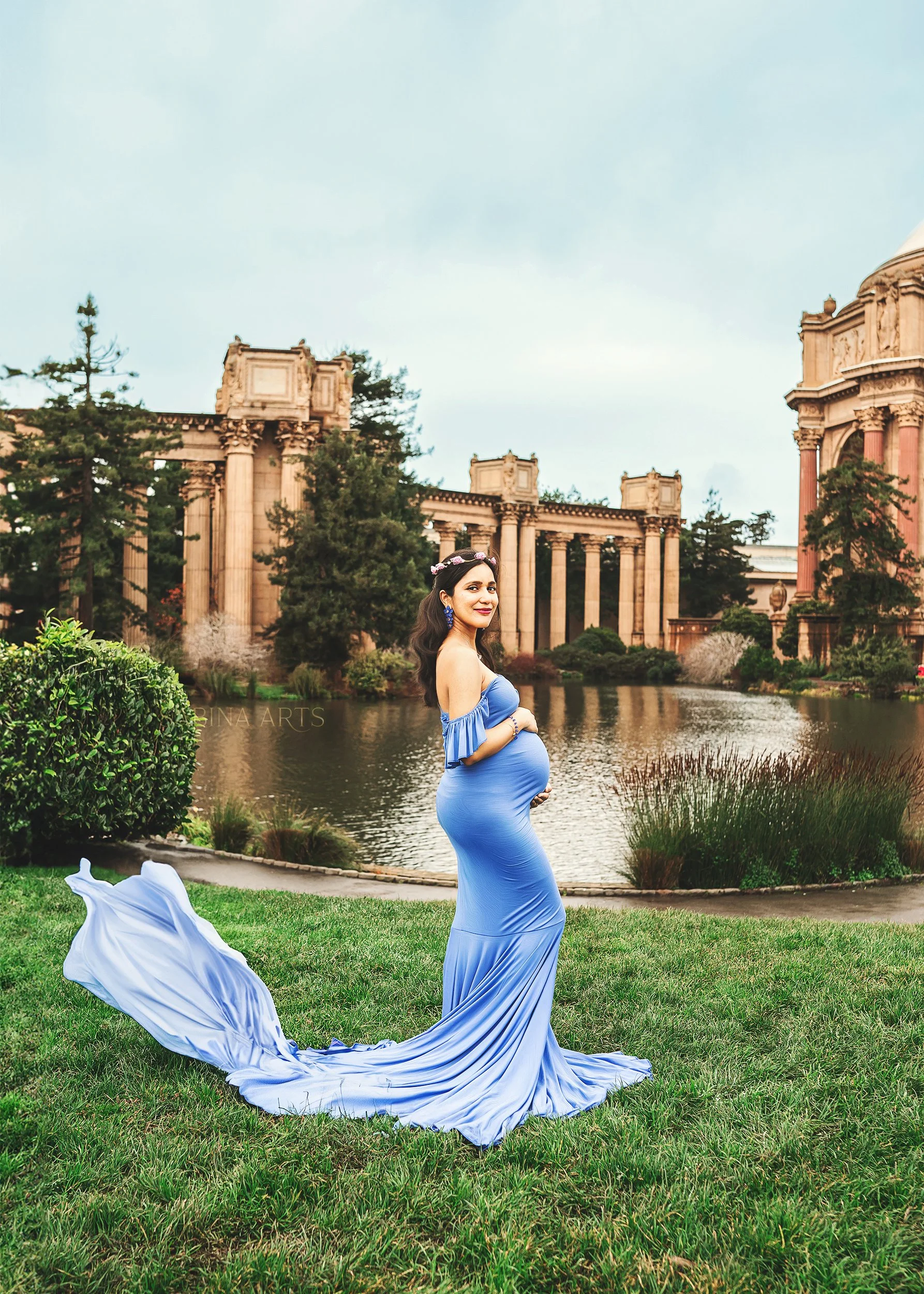 A pregnant woman in a long blue dress standing by a river in a park with historic ruins in the background.