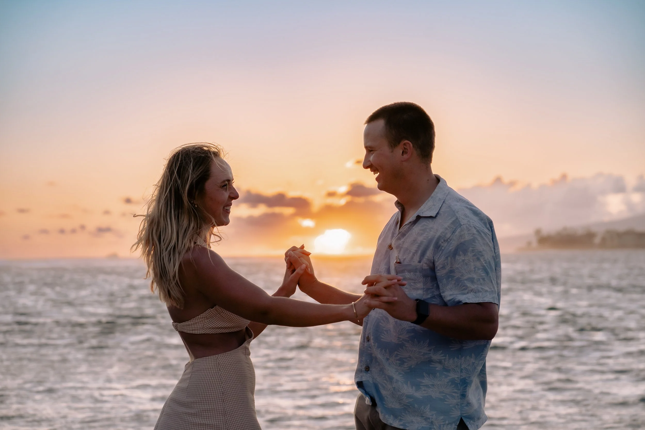 A couple dancing by the beach at sunset, smiling and holding hands.