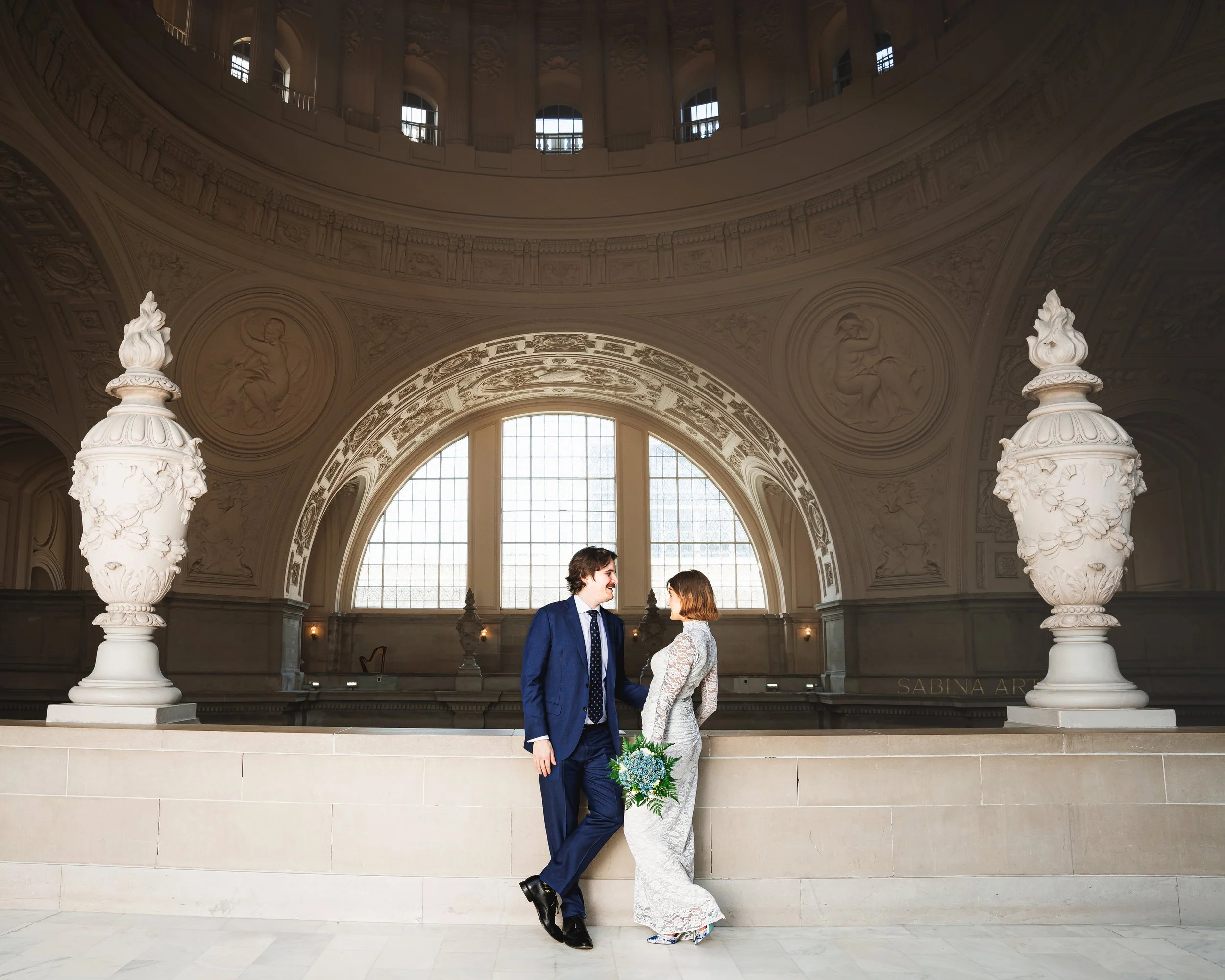 A newlywed couple standing in front of a large arched window in a grand, ornately decorated hall. The groom, dressed in a navy suit, is smiling and looking at the bride. The bride, in a white lace wedding dress, is holding a bouquet of blue flowers a