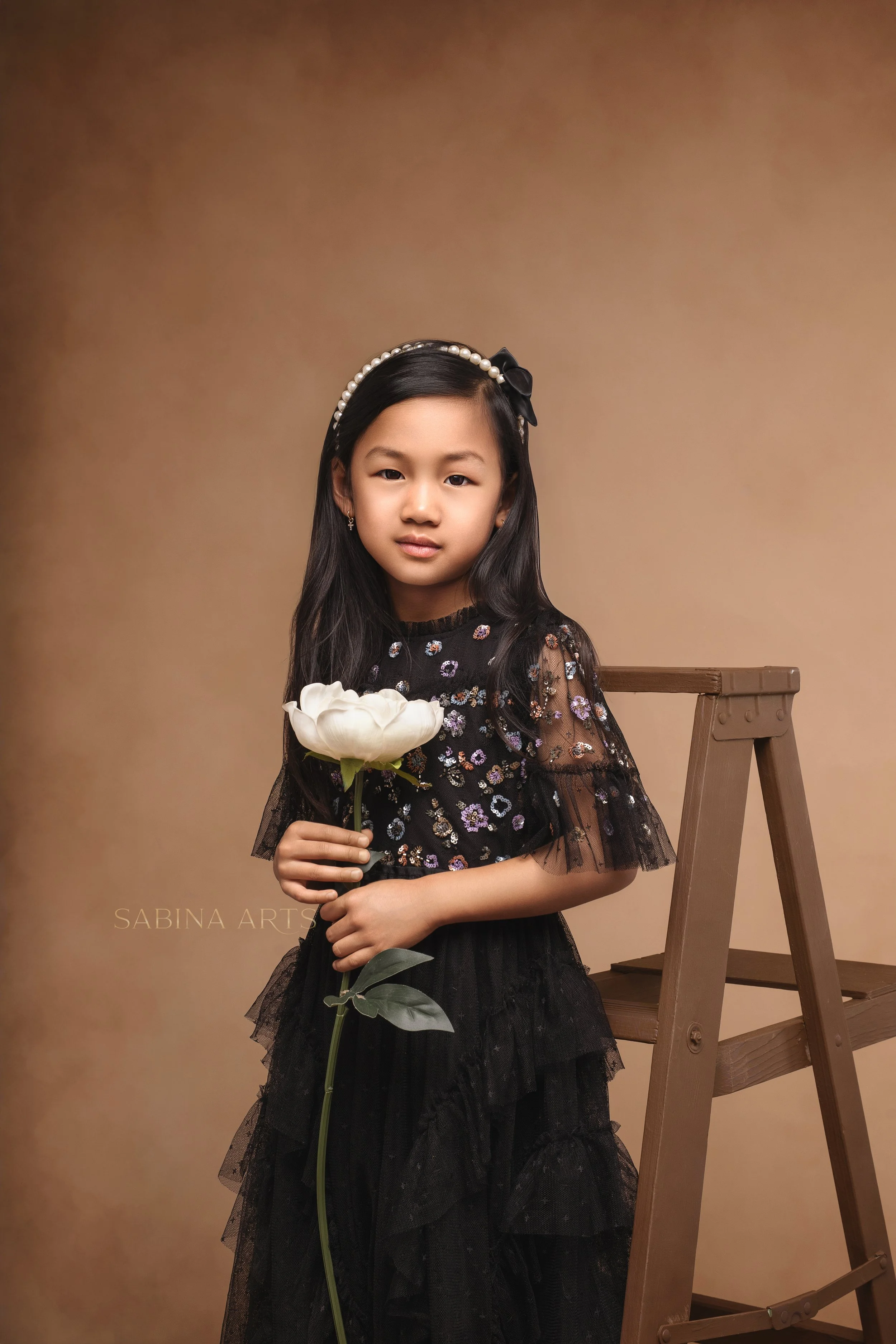 A young girl with long dark hair wearing a black dress with colorful floral embroidery, a pearl headband with a black bow, and small earrings. She is holding a white flower and standing next to a wooden step ladder against a plain brown background.