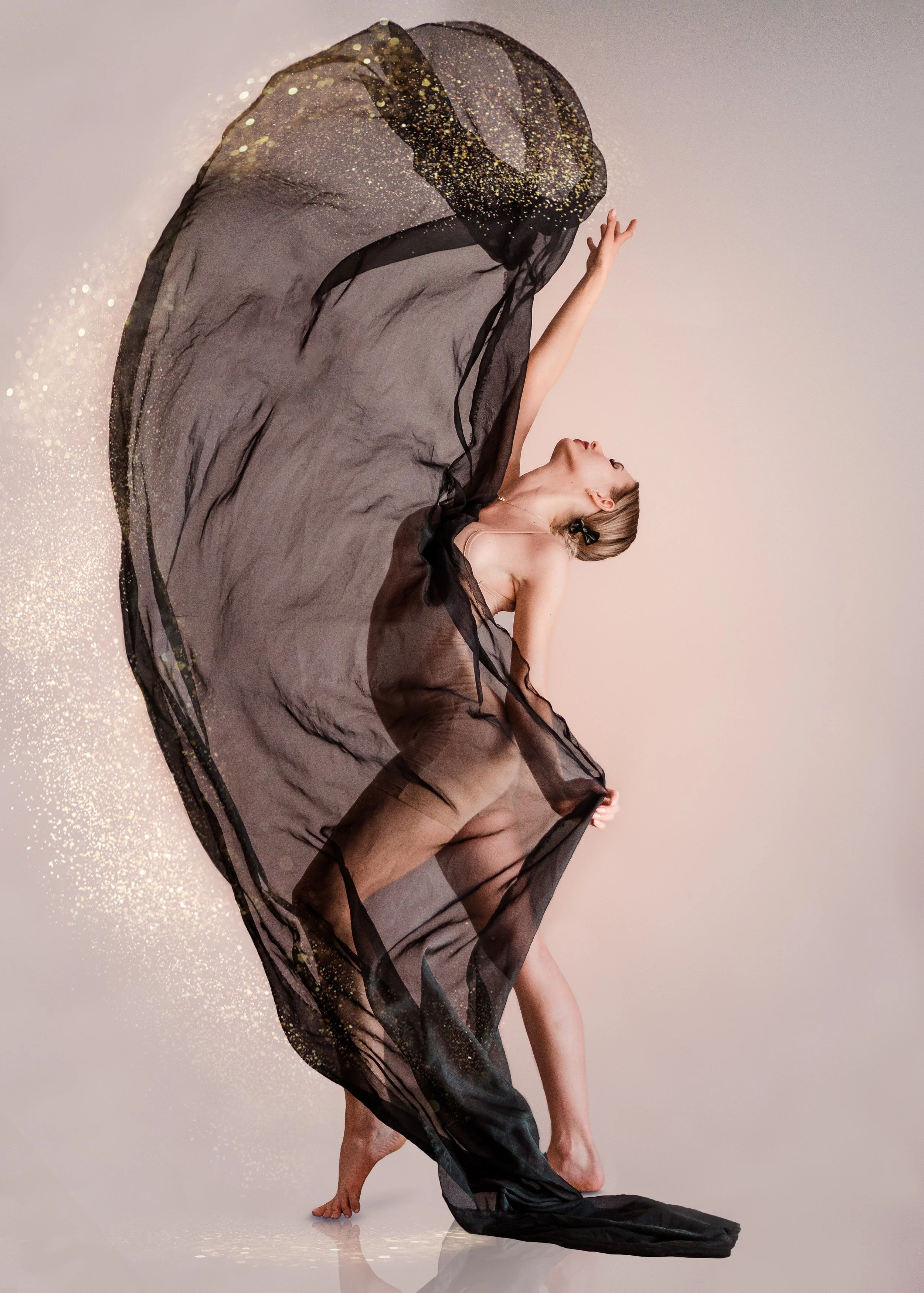 A female dancer in nude attire gracefully poses with one arm raised and her head tilted back, draped in a flowing black sheer fabric with gold accents against a plain background.