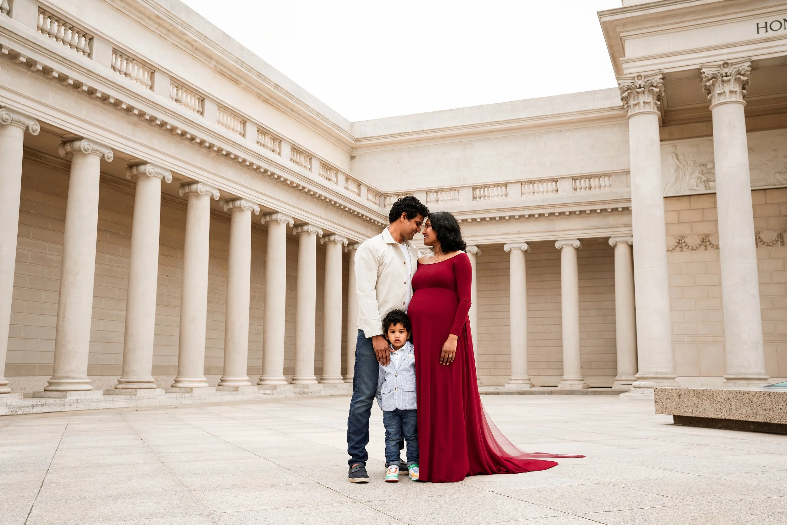 A family of four standing close together outdoors in front of a classical building with white columns. The woman is wearing a long red dress, the man is in a white shirt and jeans, and two children are with them—one in a blue jacket and jeans, and the other partially visible. The family is smiling and touching foreheads.