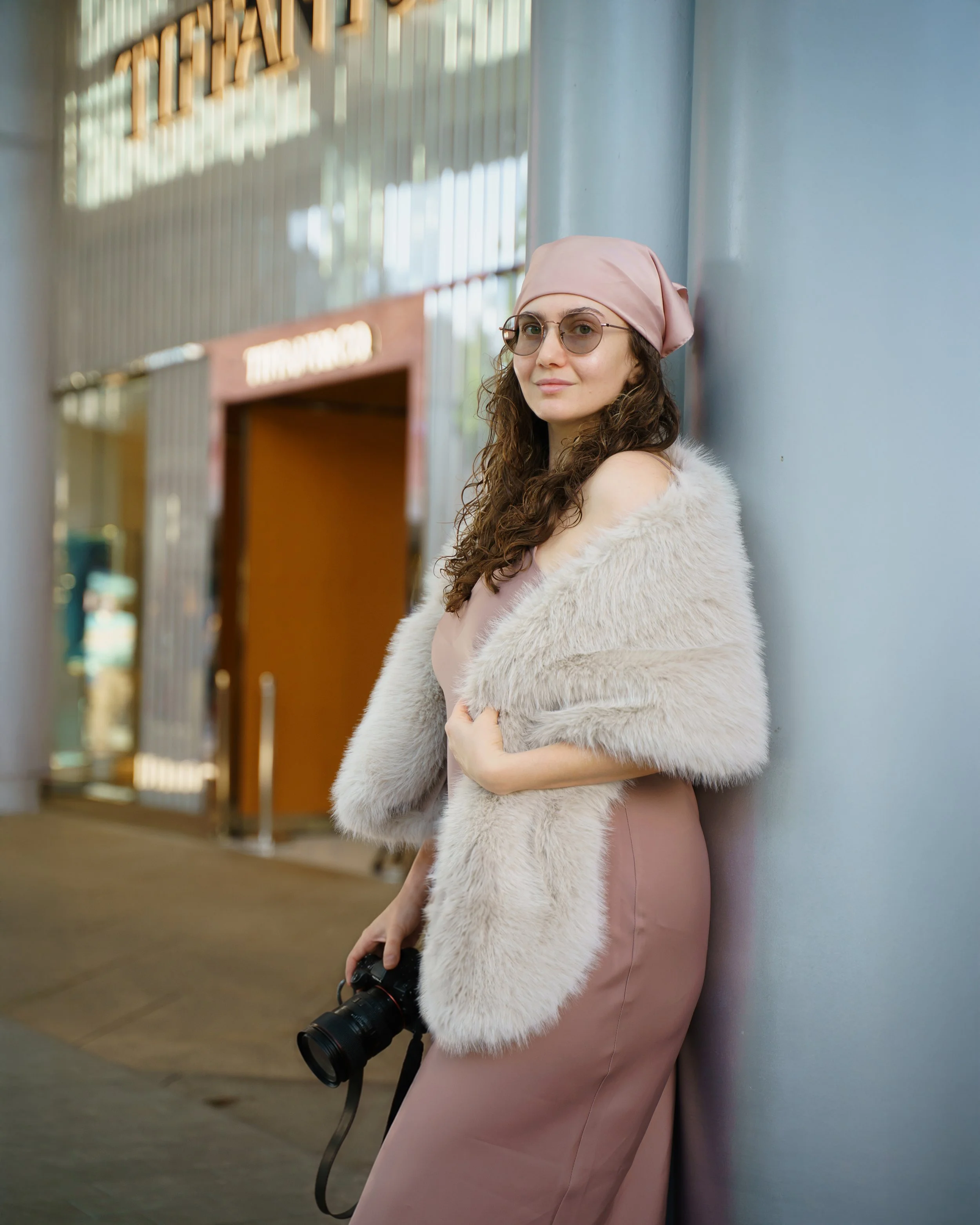A woman with curly brown hair, wearing glasses, a light pink headscarf, a white furry coat, and a pale pink dress, leaning against a light blue wall in a modern indoor setting, holding a DSLR camera with a large zoom lens.