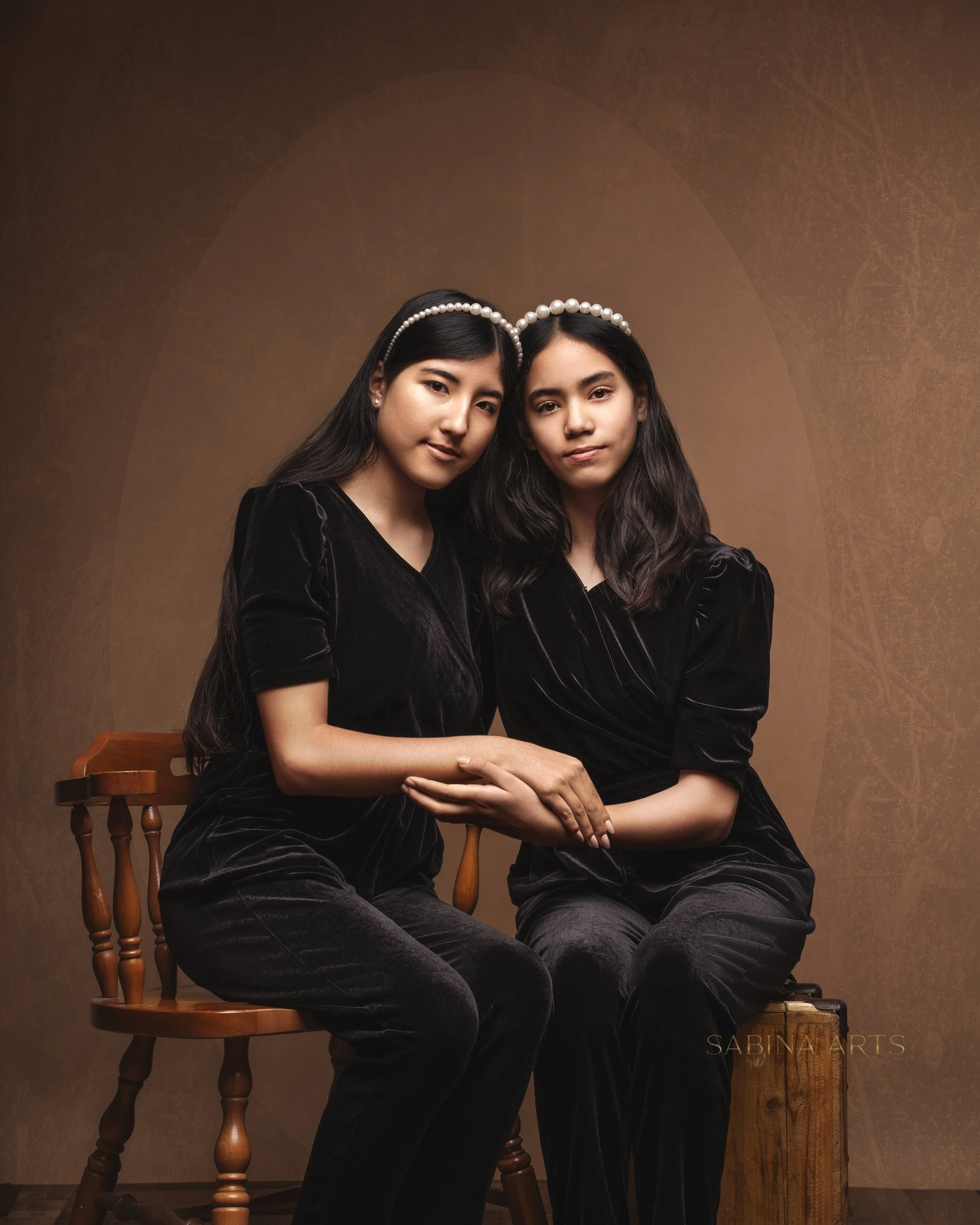 Two young women with long dark hair wearing black velvet dresses and pearl headbands, sitting close together on chairs, against a brown textured background.