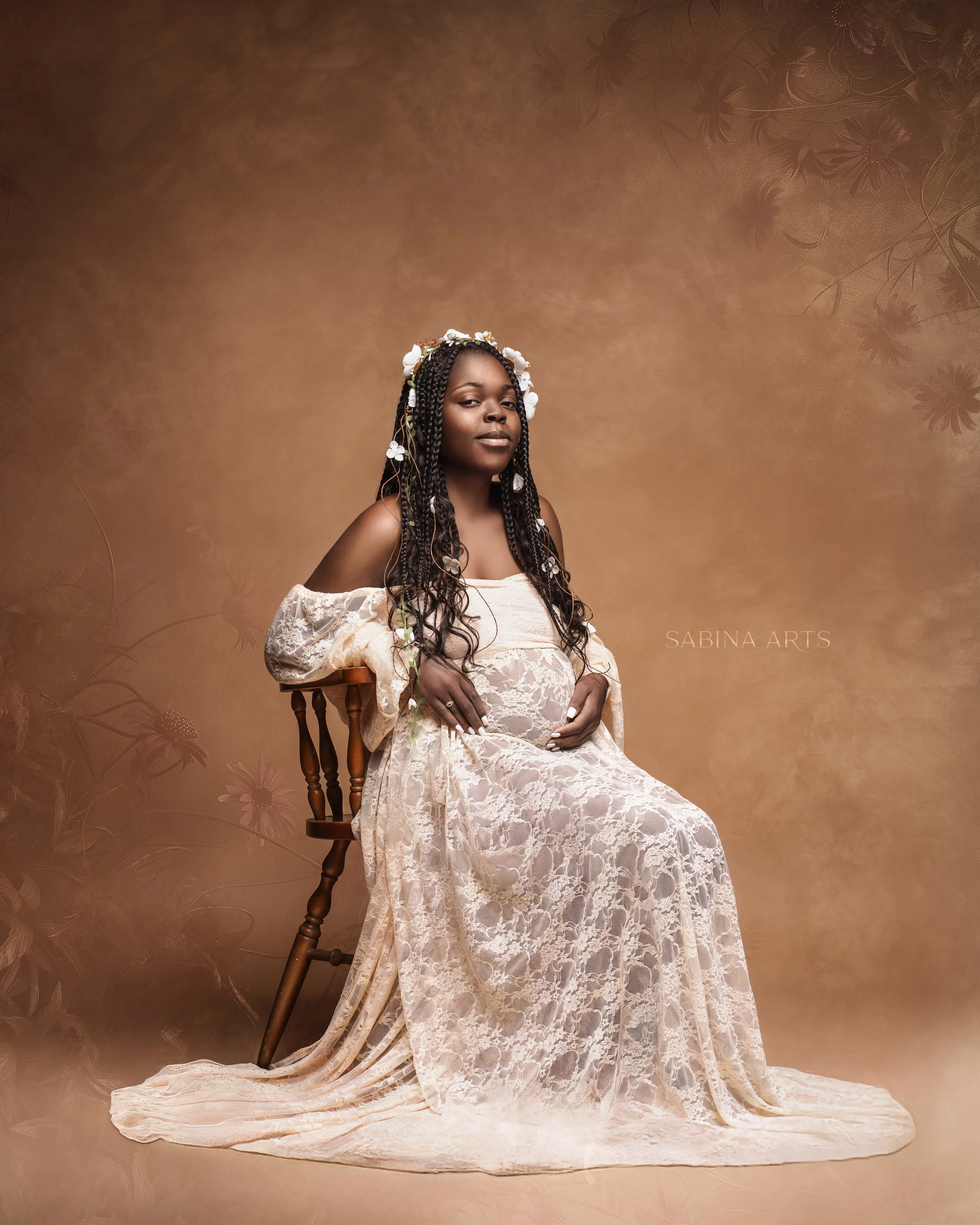 Portrait of a woman with dark skin, long braided hair with white flowers, wearing a cream lace off-shoulder dress, sitting on a wooden chair in front of a warm brown floral backdrop.