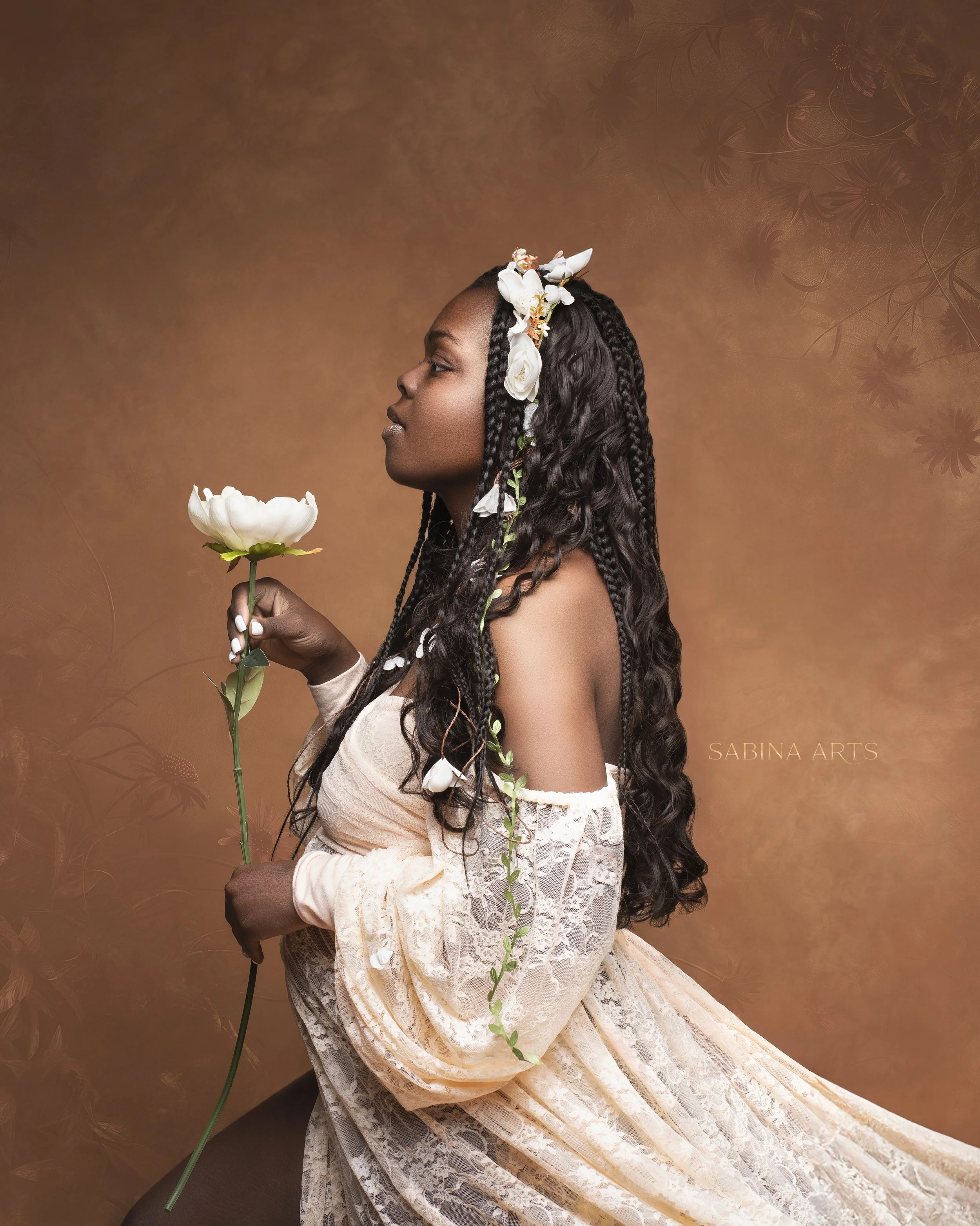 A woman with dark skin and long braided hair, wearing a light, lacy dress, holds a white flower. She has a floral headband with white flowers. The background is a warm, textured brown with a faint floral pattern. The image is artistically styled with