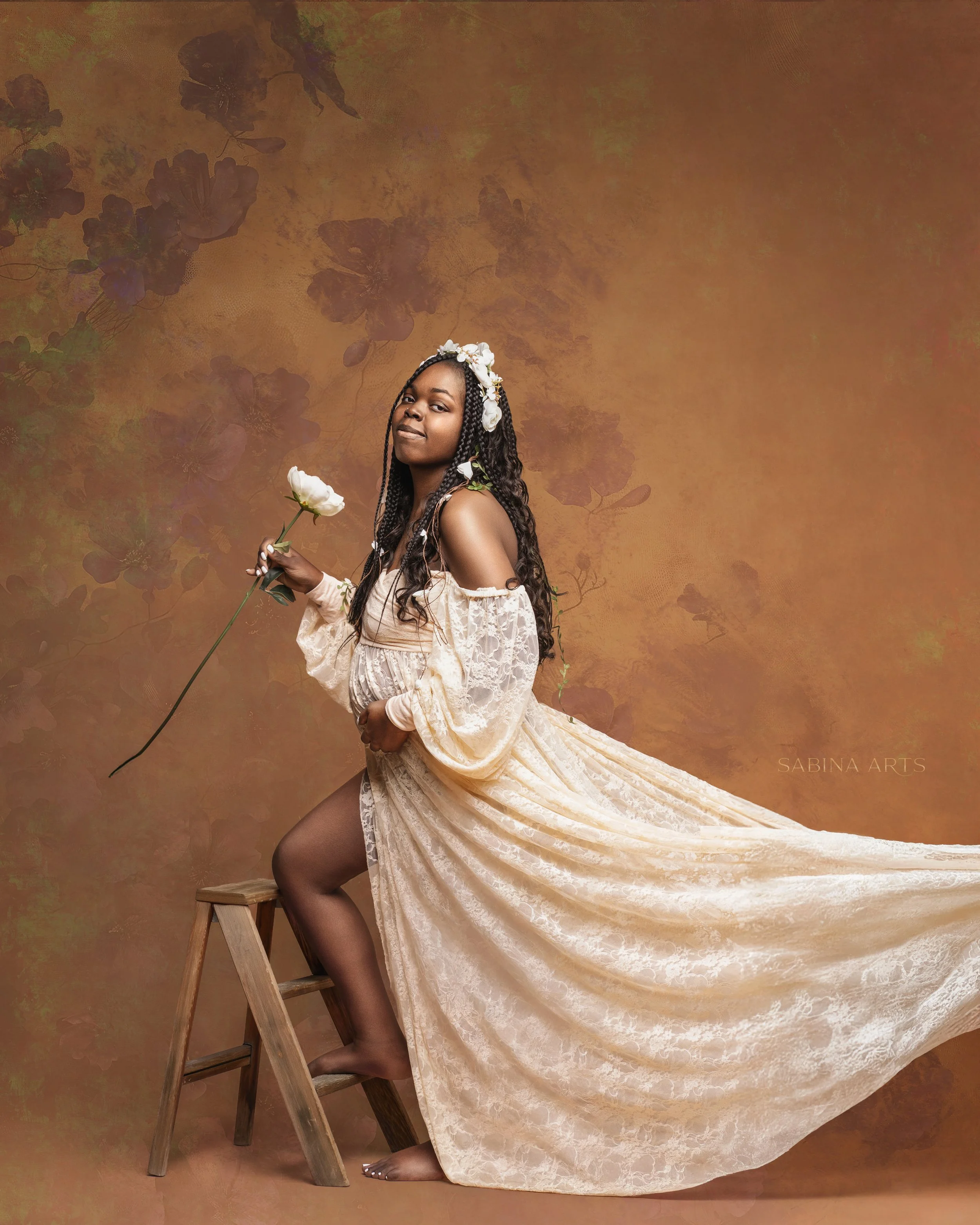 A woman with long braided hair, wearing a white floral crown and lace off-shoulder dress, holding a white flower, standing next to a wooden ladder on a light brown backdrop with floral patterns.