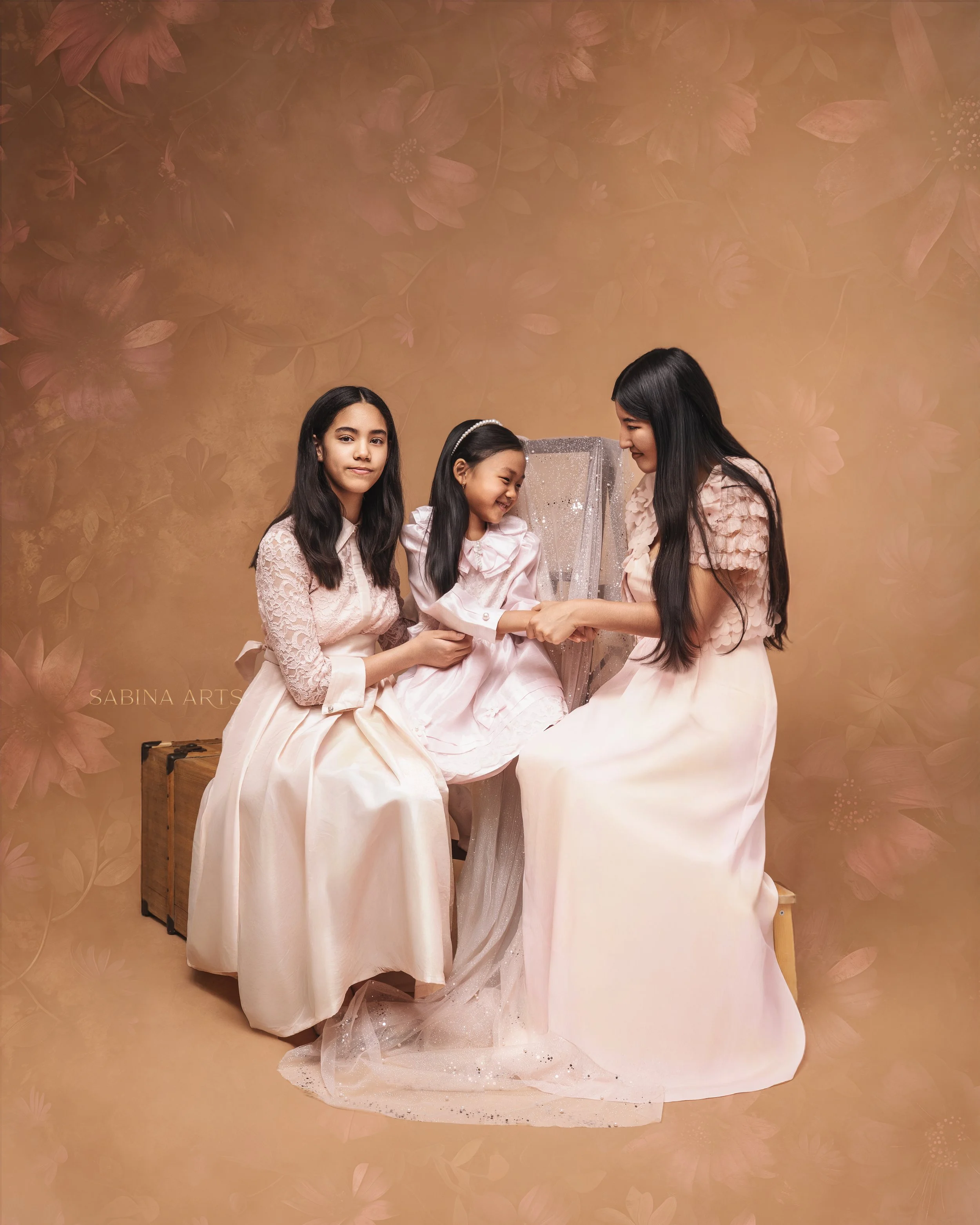 Three young girls dressed in elegant, matching pink and white dresses are gathered around a jewelry display, smiling and looking at the jewelry, with a floral patterned background.