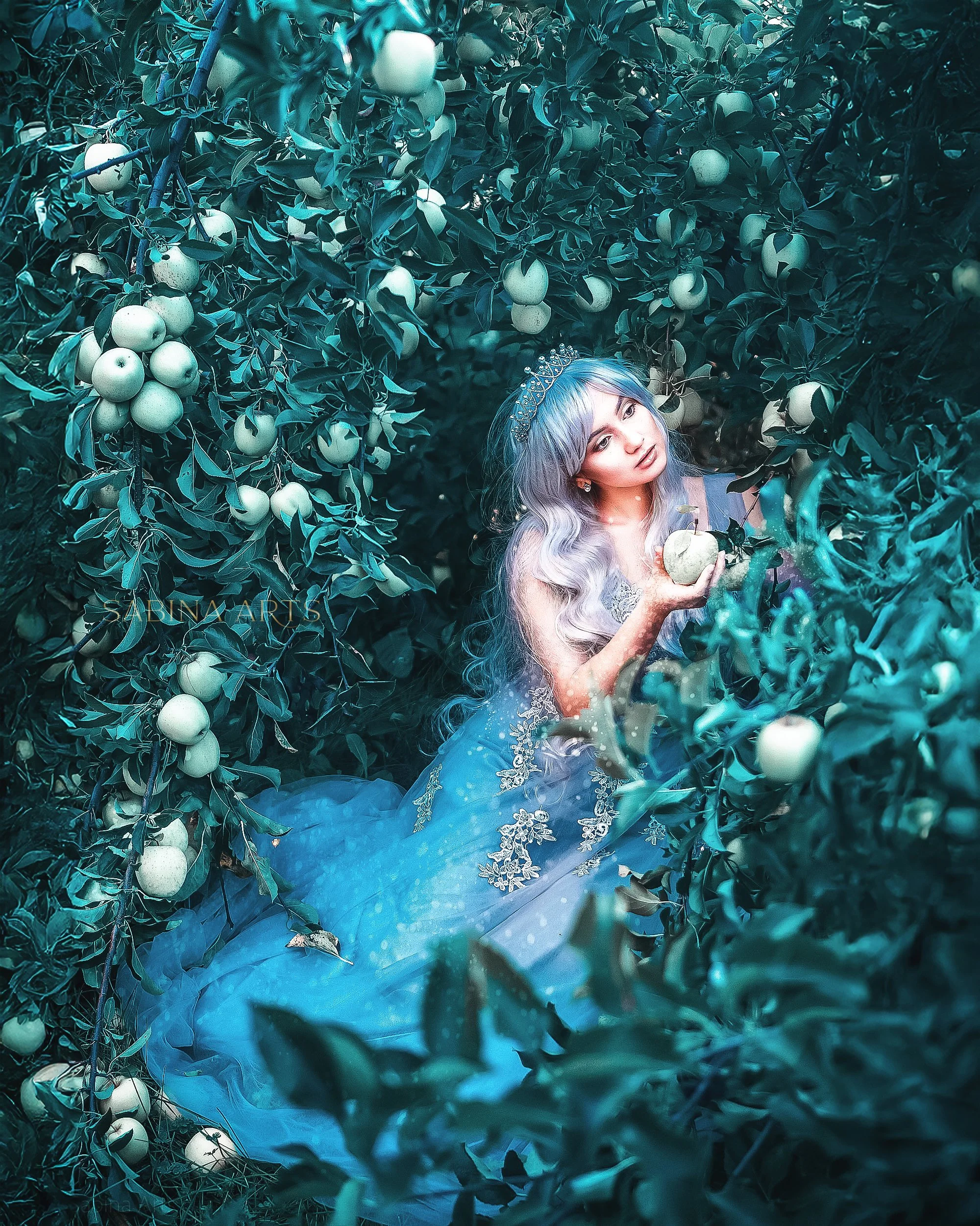 A woman with long curly silver hair, wearing a tiara and a blue dress, is standing in an orchard surrounded by green apples and leafy branches.