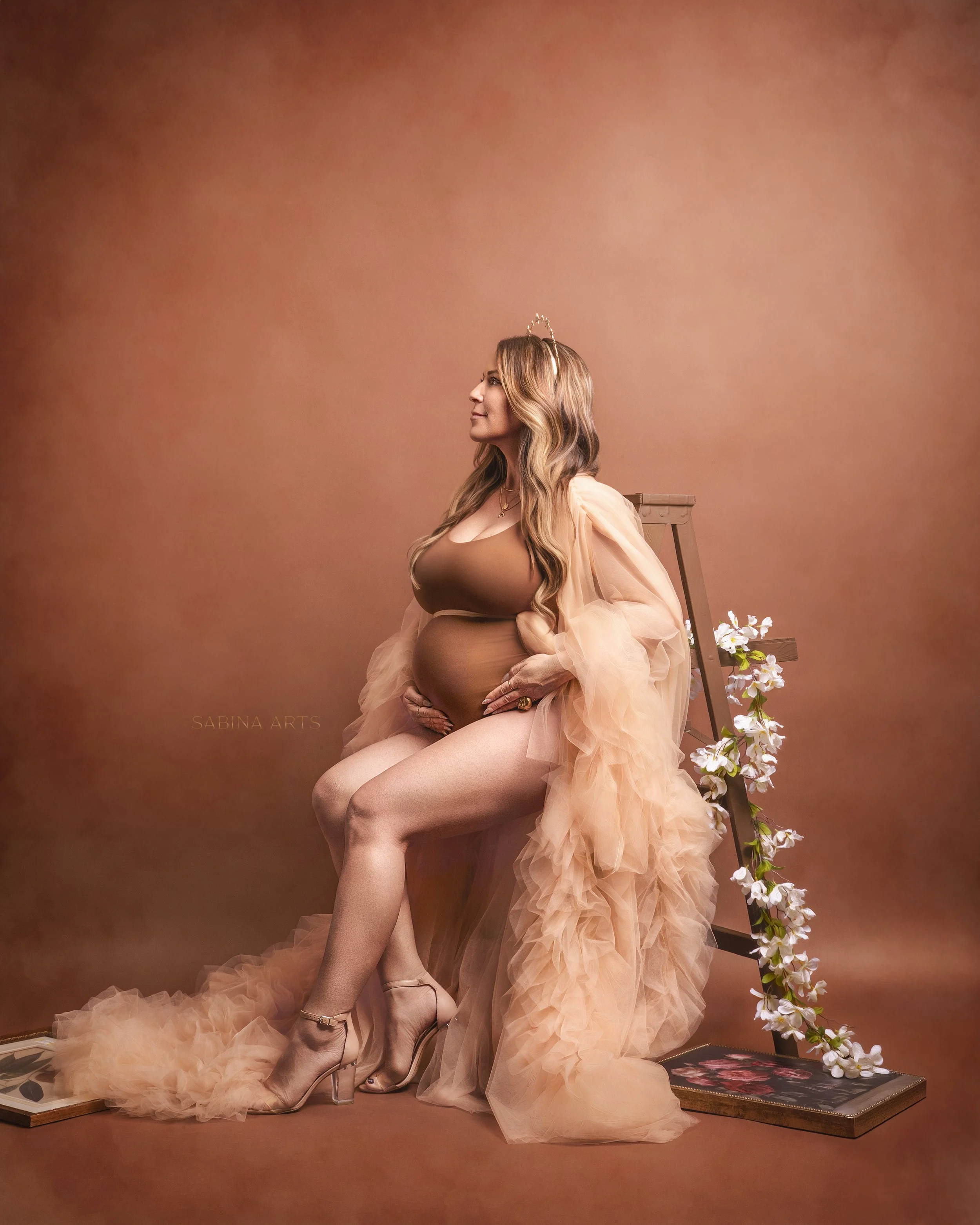 A pregnant woman with long wavy hair wearing a beige dress, a tulle shawl, and beige heels, sitting on the floor against a brown background. She has a delicate gold headband and a necklace, and is smiling while looking to her right. White flowers cas
