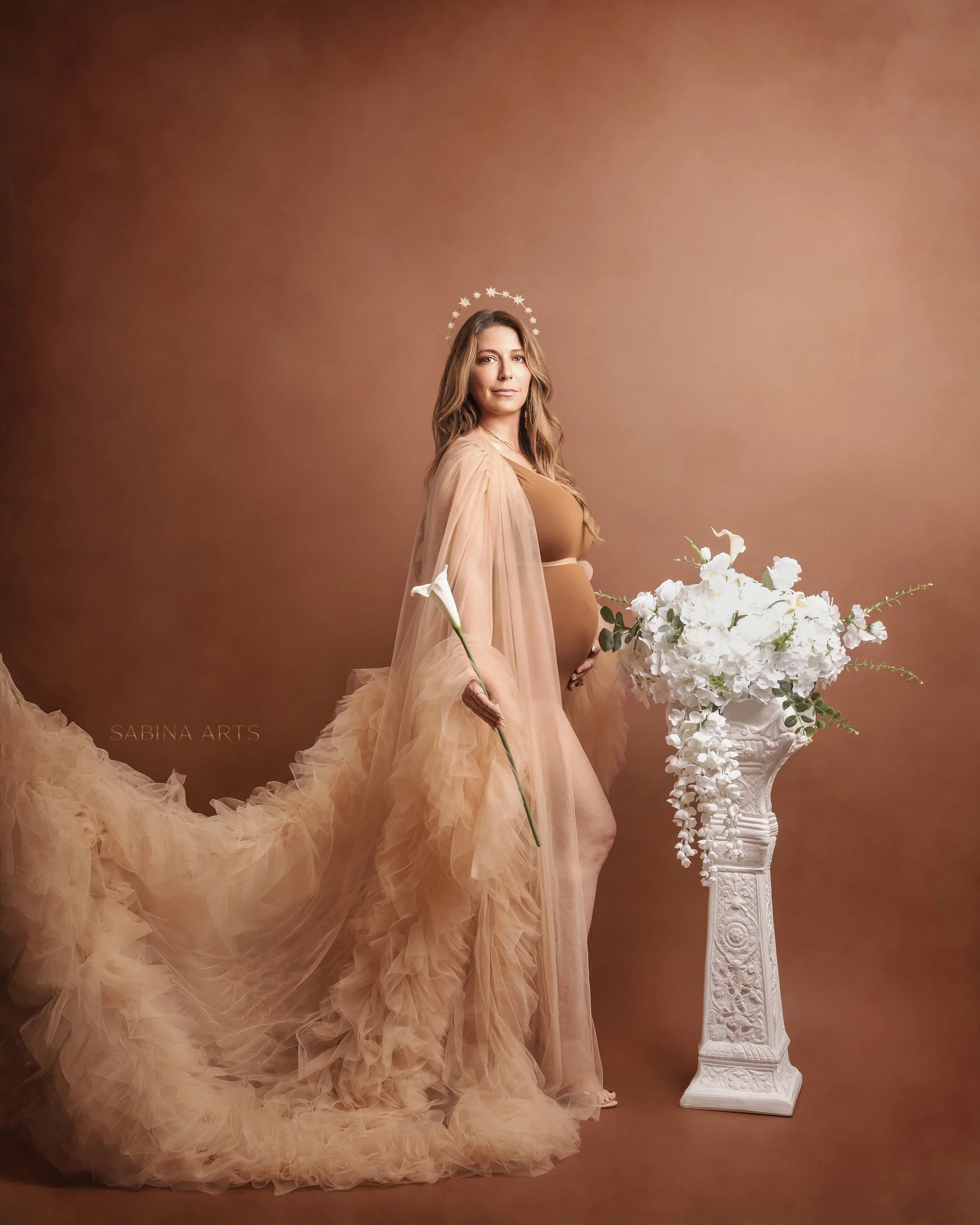 A woman with long, wavy hair dressed in a nude gown with flowing sheer fabric, standing in front of a plain brown background. She is wearing a star-shaped halo and holding a bouquet of white flowers, standing beside a white ornate pedestal with more 